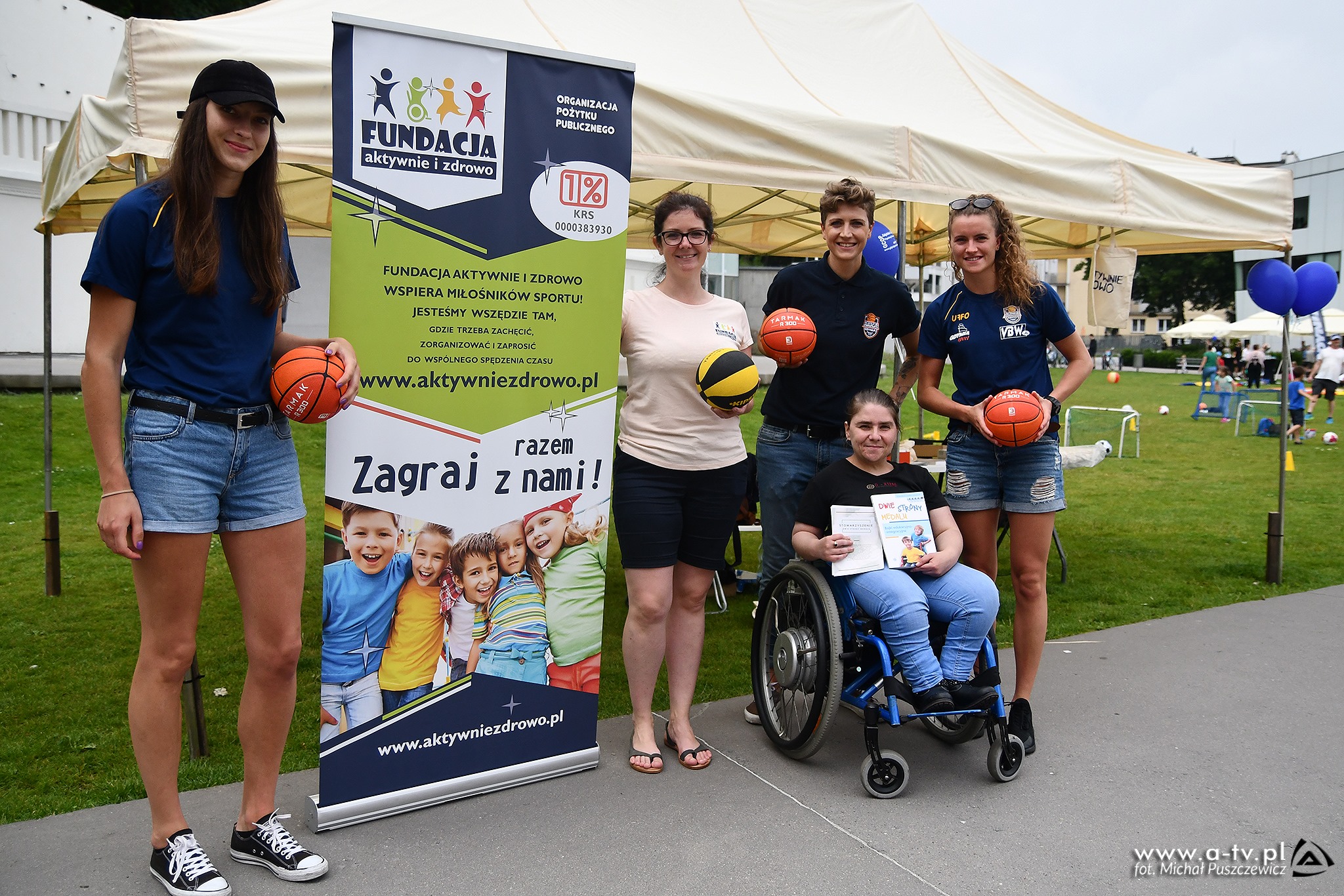 Cztery kobiety stojące z piłkami do koszykówki oraz kobieta na wózku inwalidzkim z dyplomami w ręce.  Obok nich banner organizacji pozarządowej.