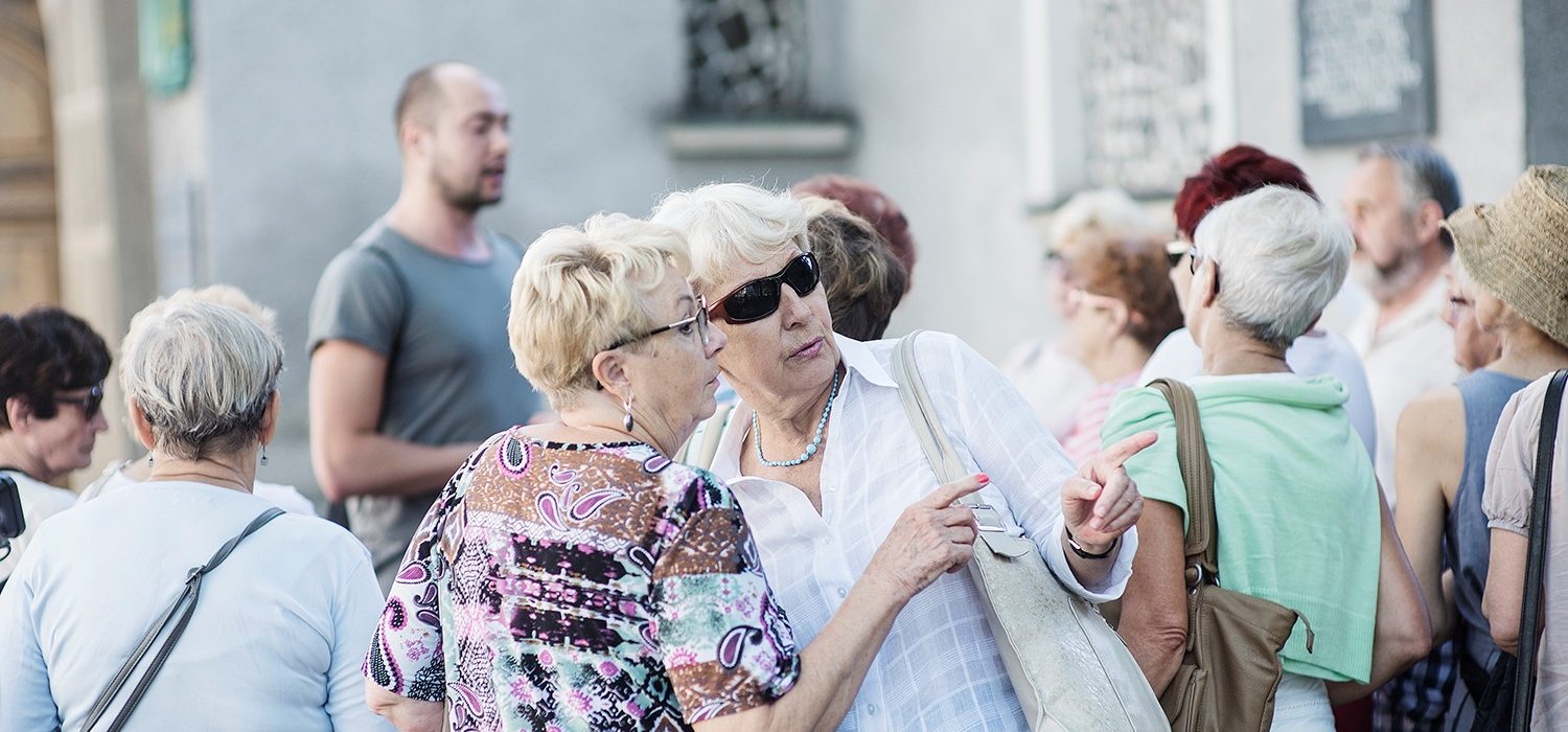 Dwie kobiety na tle tłumu ludzi. Jedna z nich posiada okulary przeciwsłoneczne. Obracają się w prawą stronę, wskazując przy tym dłonią.. 