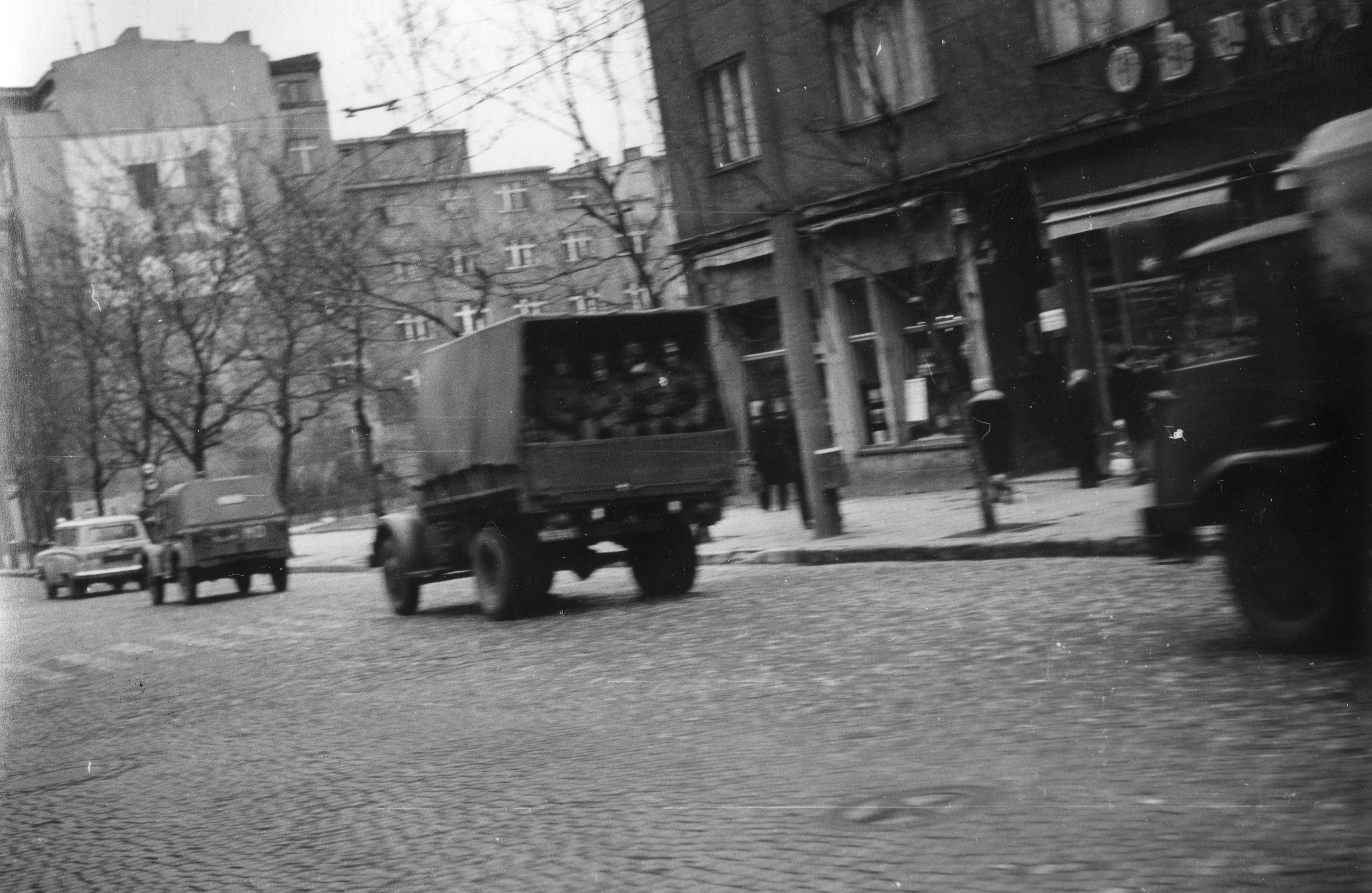 Kolumna samochodów milicyjnych. Na ciężarówkach widoczni są milicjanci. Pierwszy z jadących samochodów to "Wołga"  kombi, przypuszczalnie jest to auto widoczne na zdjęciu o sygnaturze IPN Gd 003/14 t. 88 k. 1/46. Zdjęcie wykonane na wysokości Pomnika Harcerzy w Gdyni na ul.Świętojańskiej // źródło: IPN