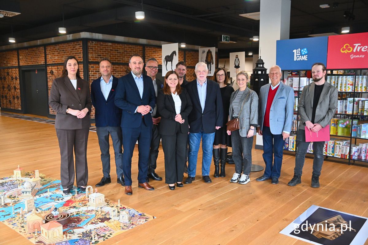 Grupa osób stojących w sali pełnej puzzli oraz makiet budynków