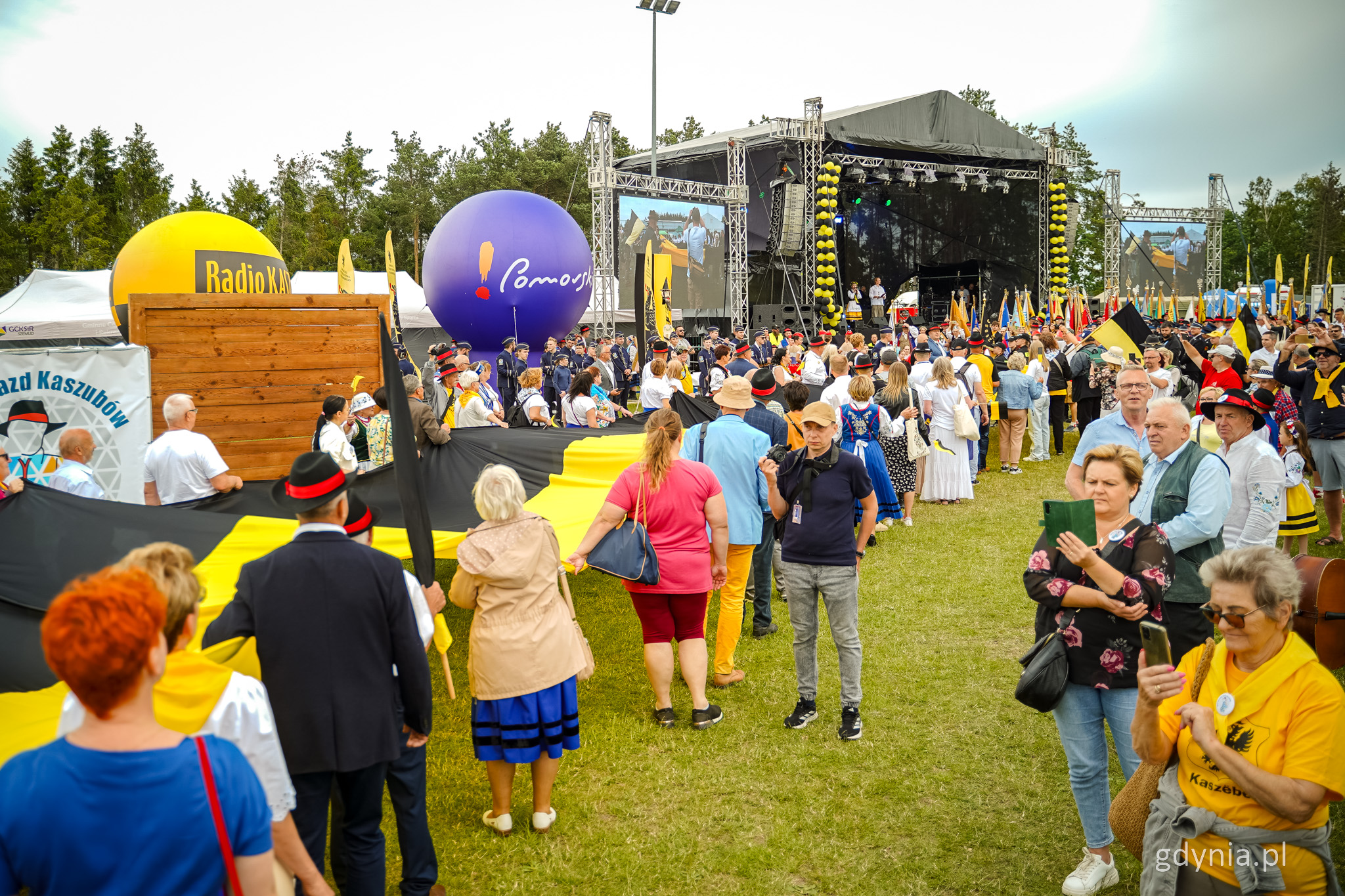 Stadion w Szemudzie, scena, teren zjazdu Kaszubów. Grupa osób stoi i trzyma czarno-żółtą kaszubską flagę