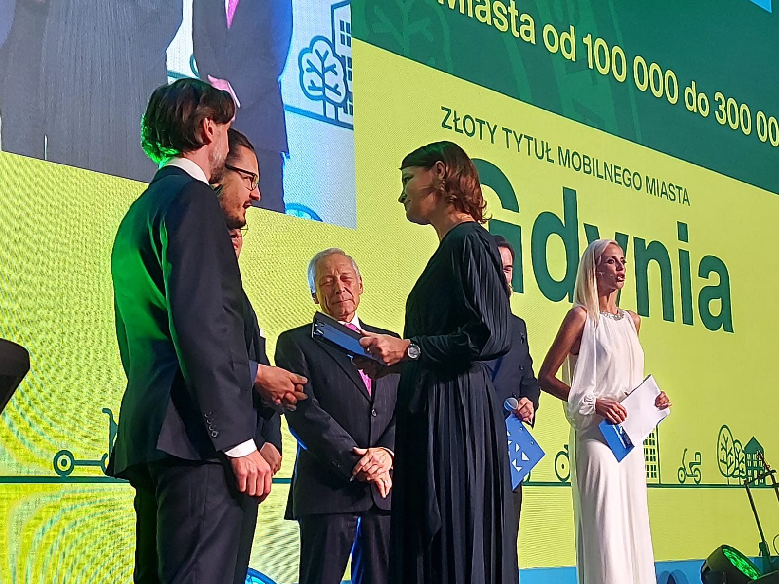 Scena Kongresu Nowej Mobilności, żółte tło z napisem "Gdynia", Katarzyna Gruszecka-Spychała odbiera nagrodę, na scenie kilka osób