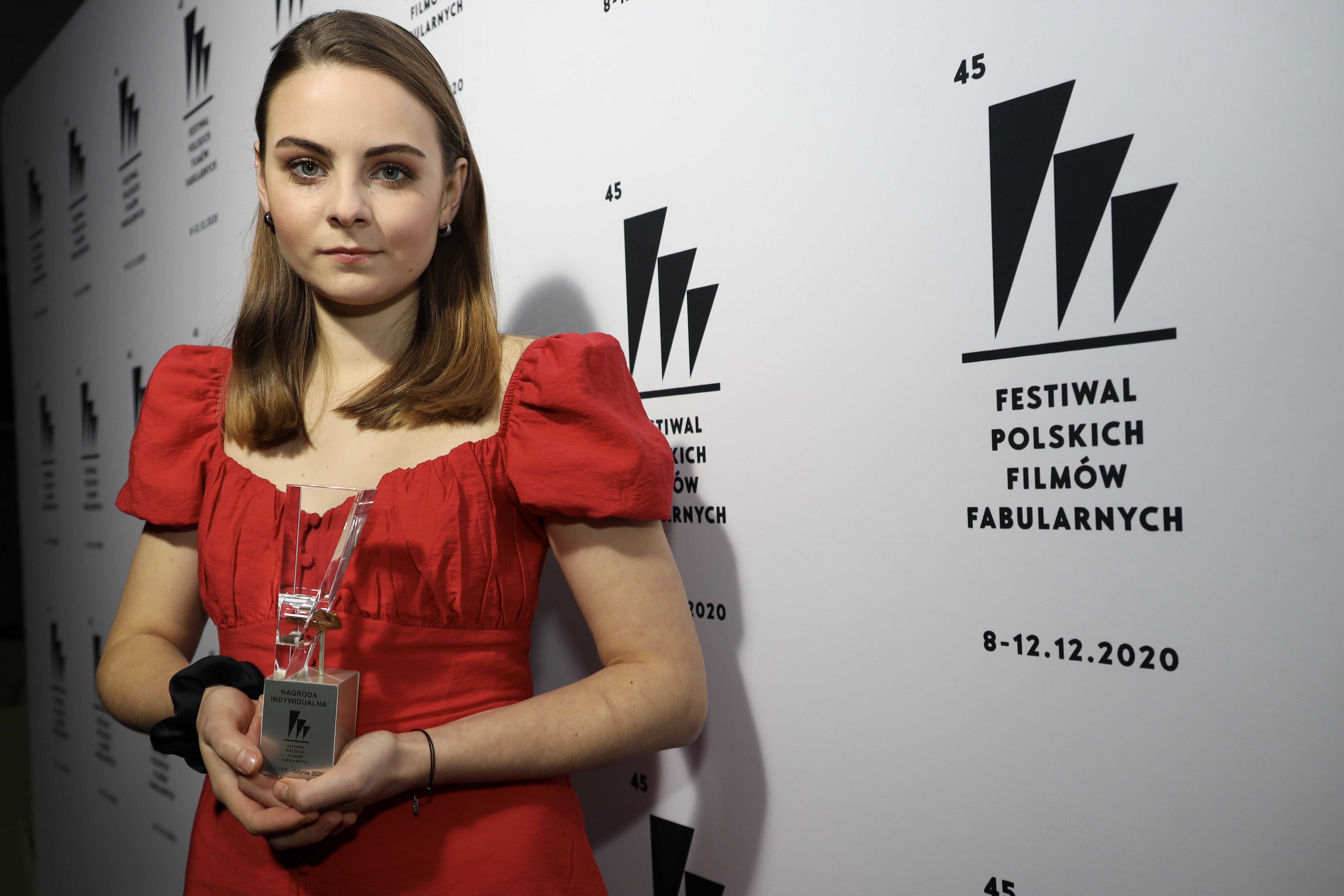 Zofia Stafiej - laureatka nagrody za debiut aktorski ze statuetką w ręku stoi na tle białej ścianki z czarnymi miniaturkami logo festiwalu // fot. Borys Skrzyński