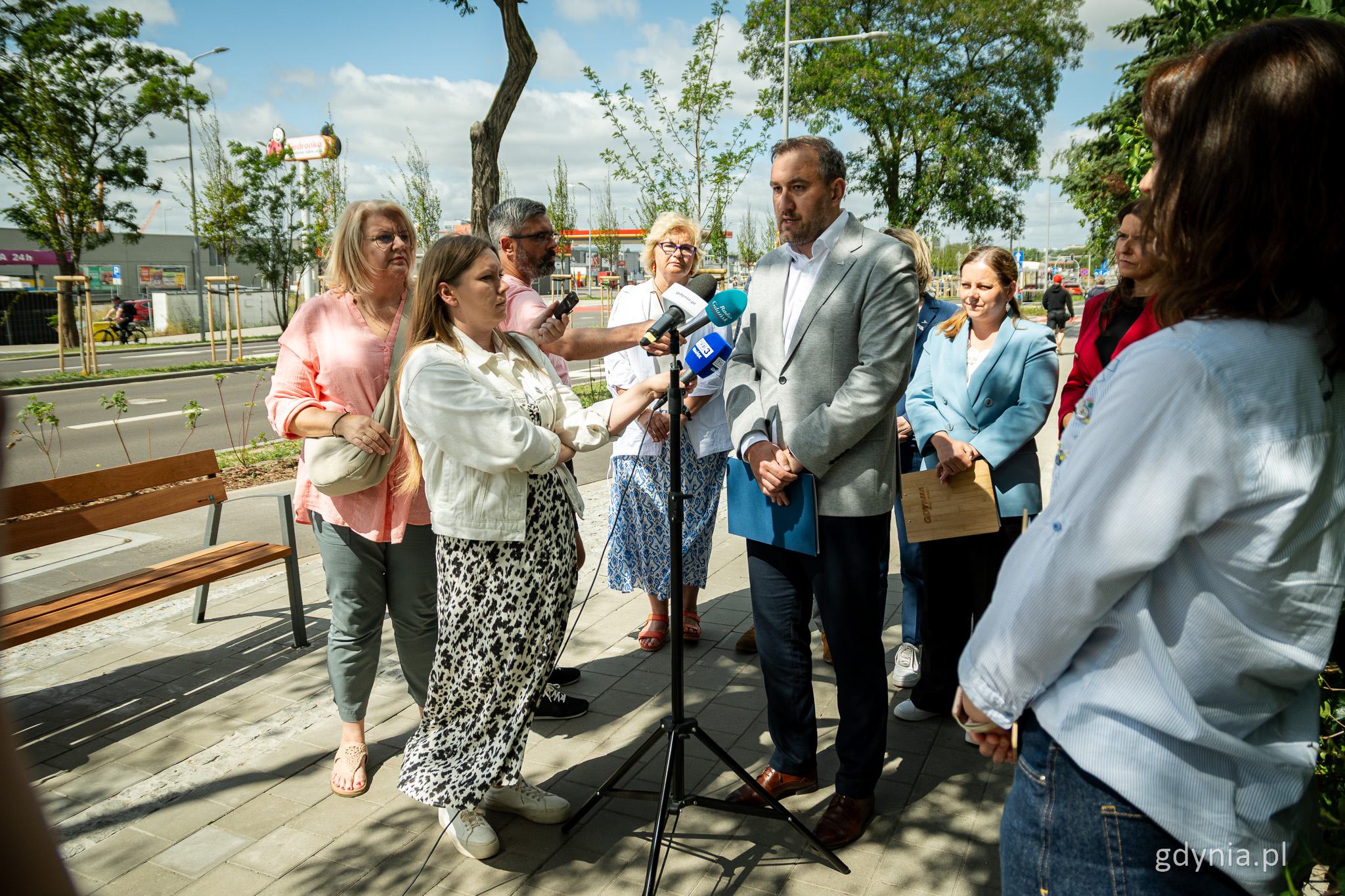 Chodnik przy ul. Unruga, słoneczny dzień, przemawia do mikrofonów Tomasz Augustyniak, obok grupa dziennikarzy