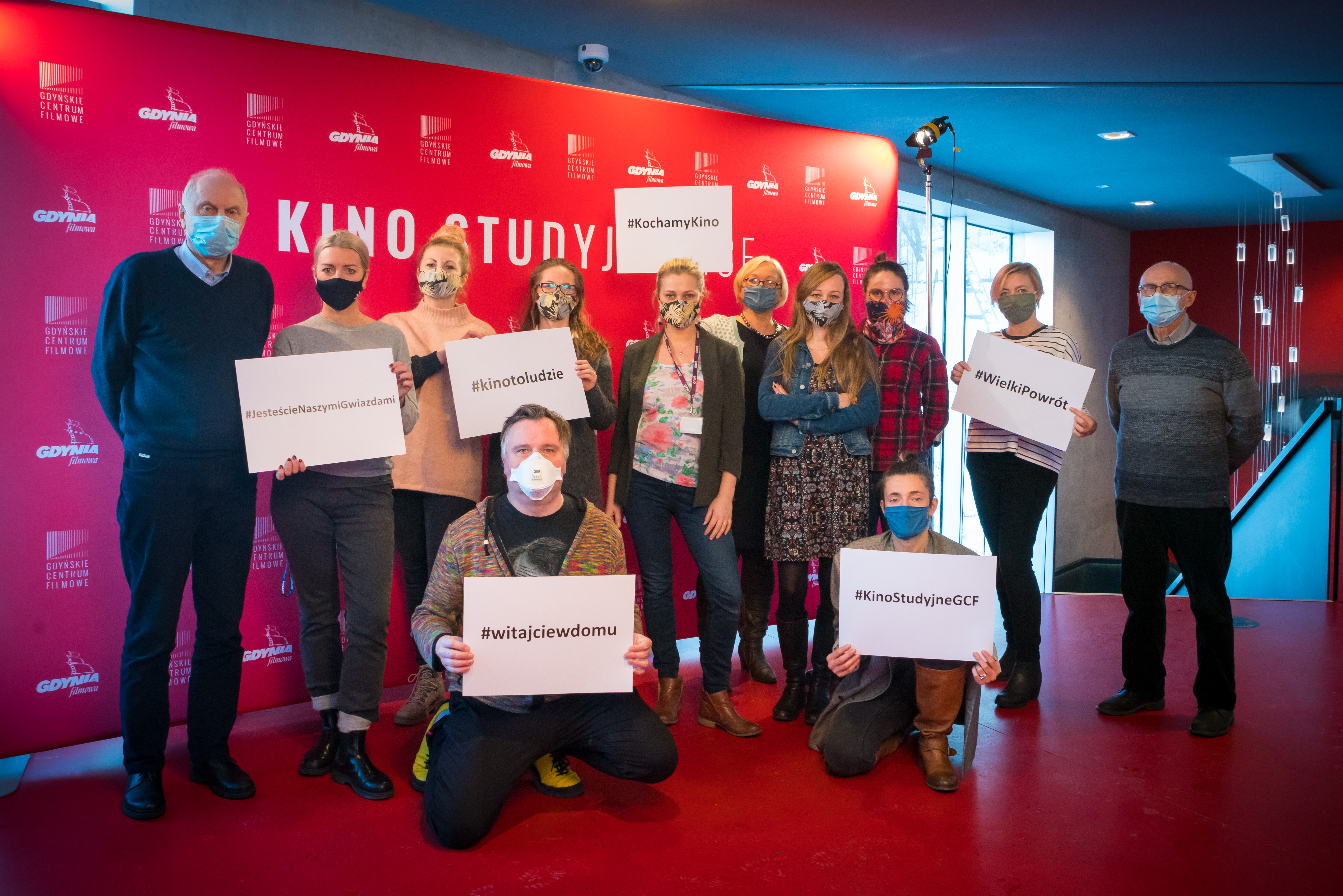 Grupa 12. osób zebrała się przed ścianką Gdyńskiego Centrum Filmowego. Osoby na zdjęciu mają założone maseczki, a niektóre osoby trzymają kartki z napisami "#witajciewdomu", "#kinotoludzie", "#kochamykino". // fot. Anna Rezulak / KFP