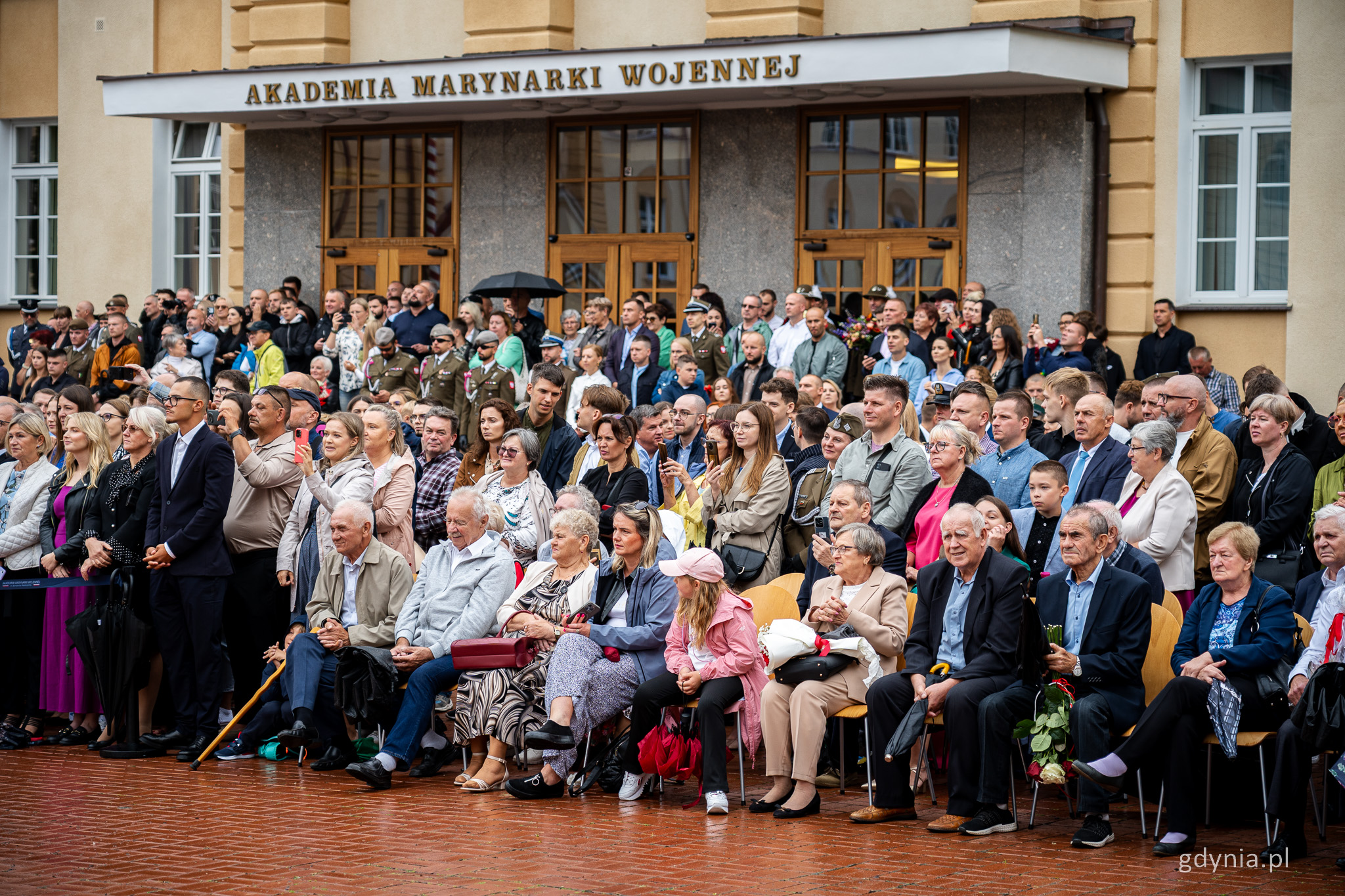 Plac apelowy AMW, grupa osób siedzi i stoi, przyglądając się promocjom oficerskim, w tle budynki