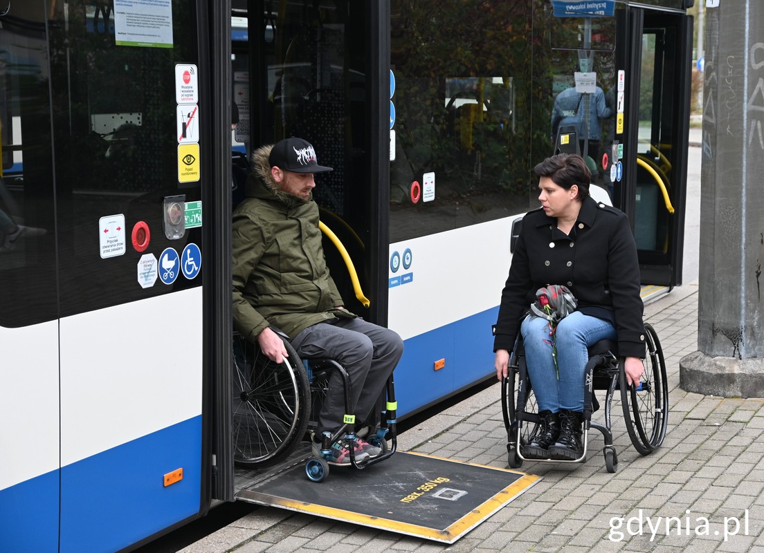 Dwie osoby na wózkach: jedna wyjeżdża z autobusu, druga czeka przed drzwiami