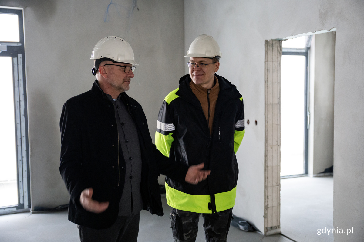 Dwaj mężczyźni w kaskach rozmawiają wewnątrz budynku na budowie.