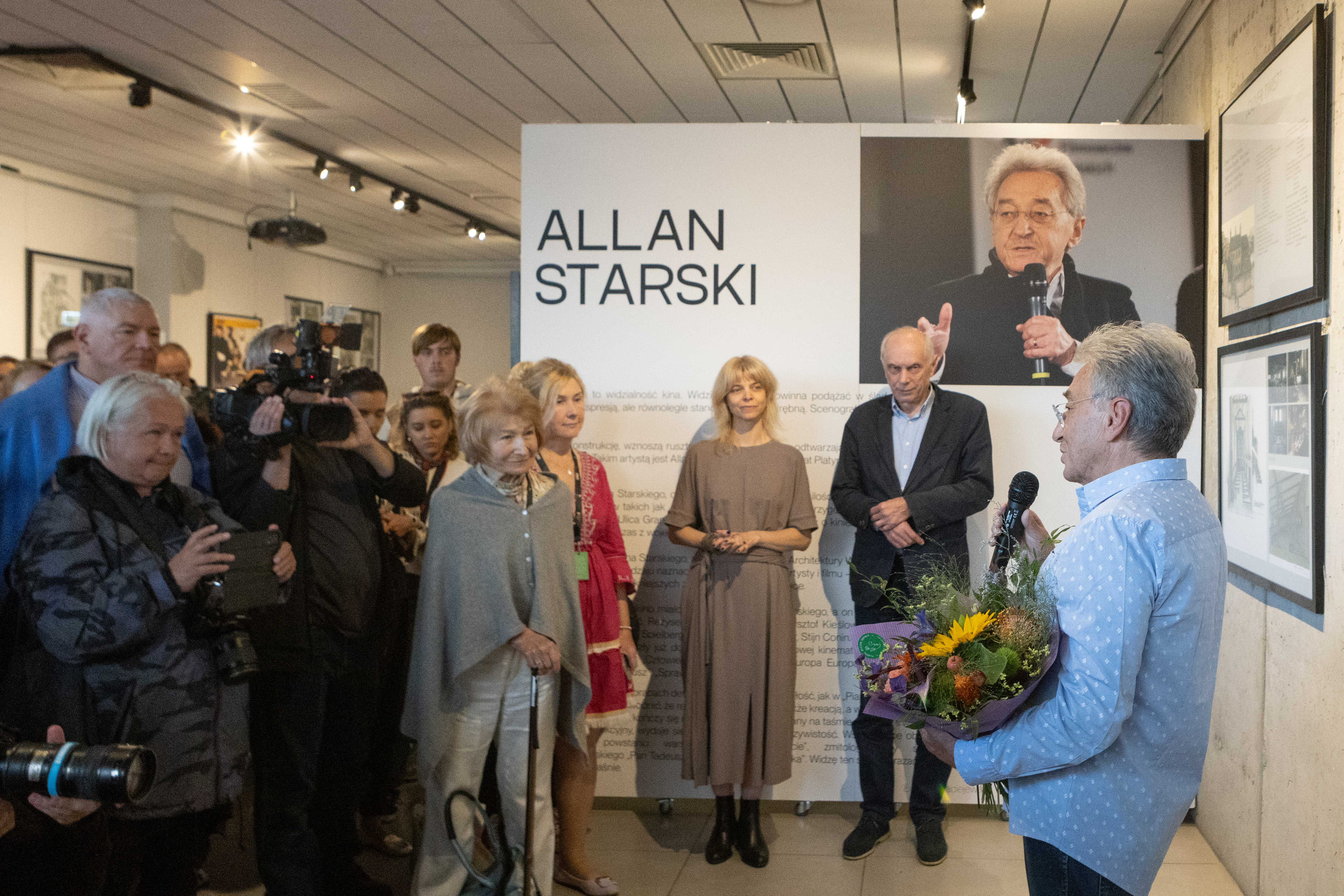 Galeria GCF, wystawa Allana Starskiego, Allan Starski odbiera kwiaty i przemawia, obok tłum ludzi, na ścianach ekspozycja w ramach