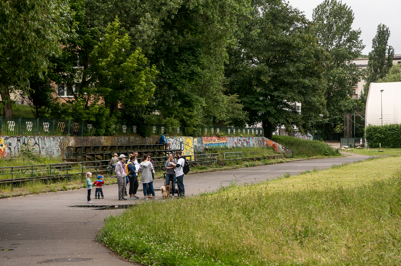 Dawne boisko przy al. Piłsudskiego, zieleń, trawa, drzewa, grupa ludzi rozmawia na alejce
