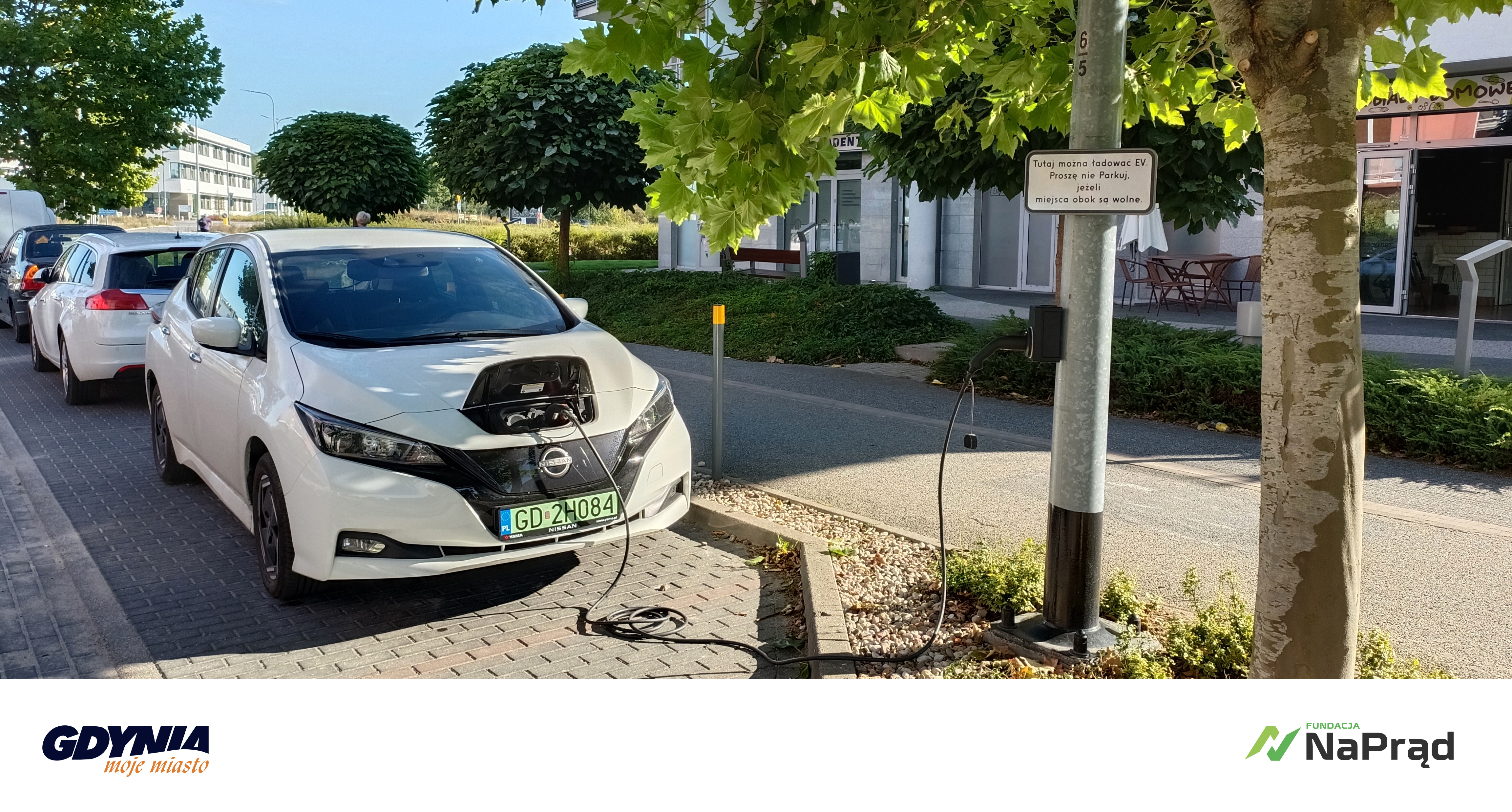 „Electric Street” - wspólny projekt miasta Gdynia i Fundacji #NaPrąd. Zdjęcie samochodu elektrycznego w zatoczce, podłączony kablem do słupa oświetleniowego. Obok inne samochody, ulica, budynki