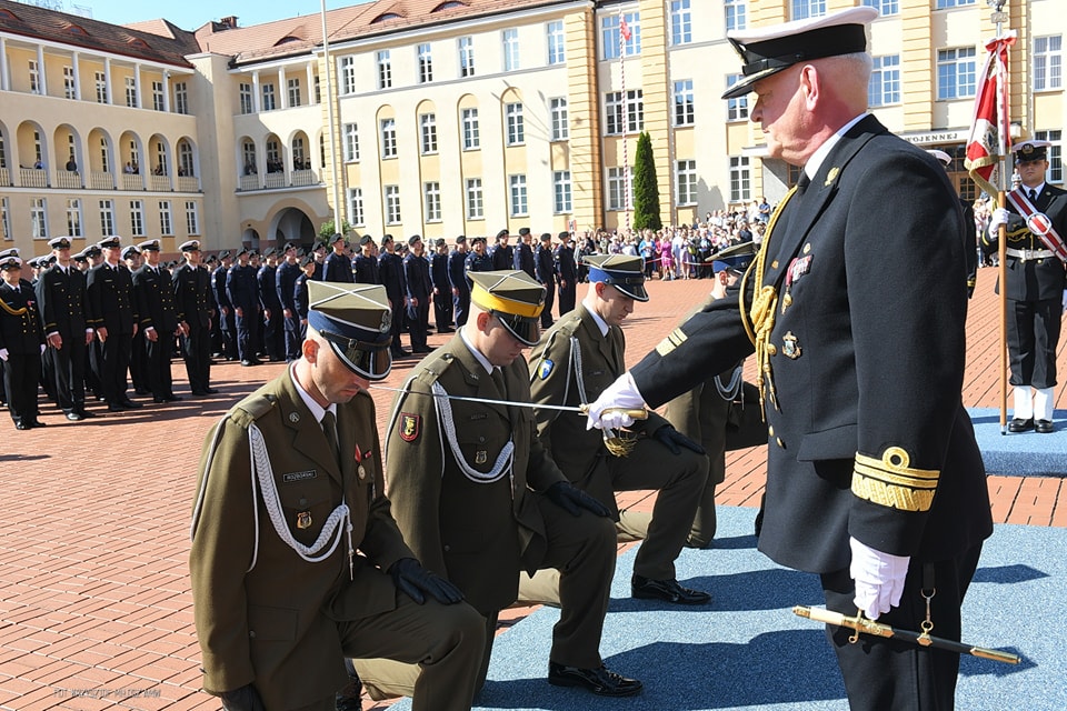 Akademia Marynarki Wojennej, plac, w tle budynek, uroczystość promocji oficerskiej, klęczą oficerowie, dowódca dotyka pałaszem ramienia jednego z nich, w tle żołnierze