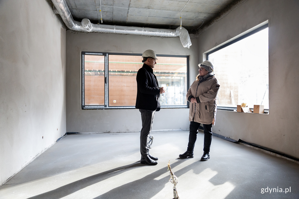 Kobieta i mężczyzna w kaskach rozmawiają wewnątrz budynku na budowie.