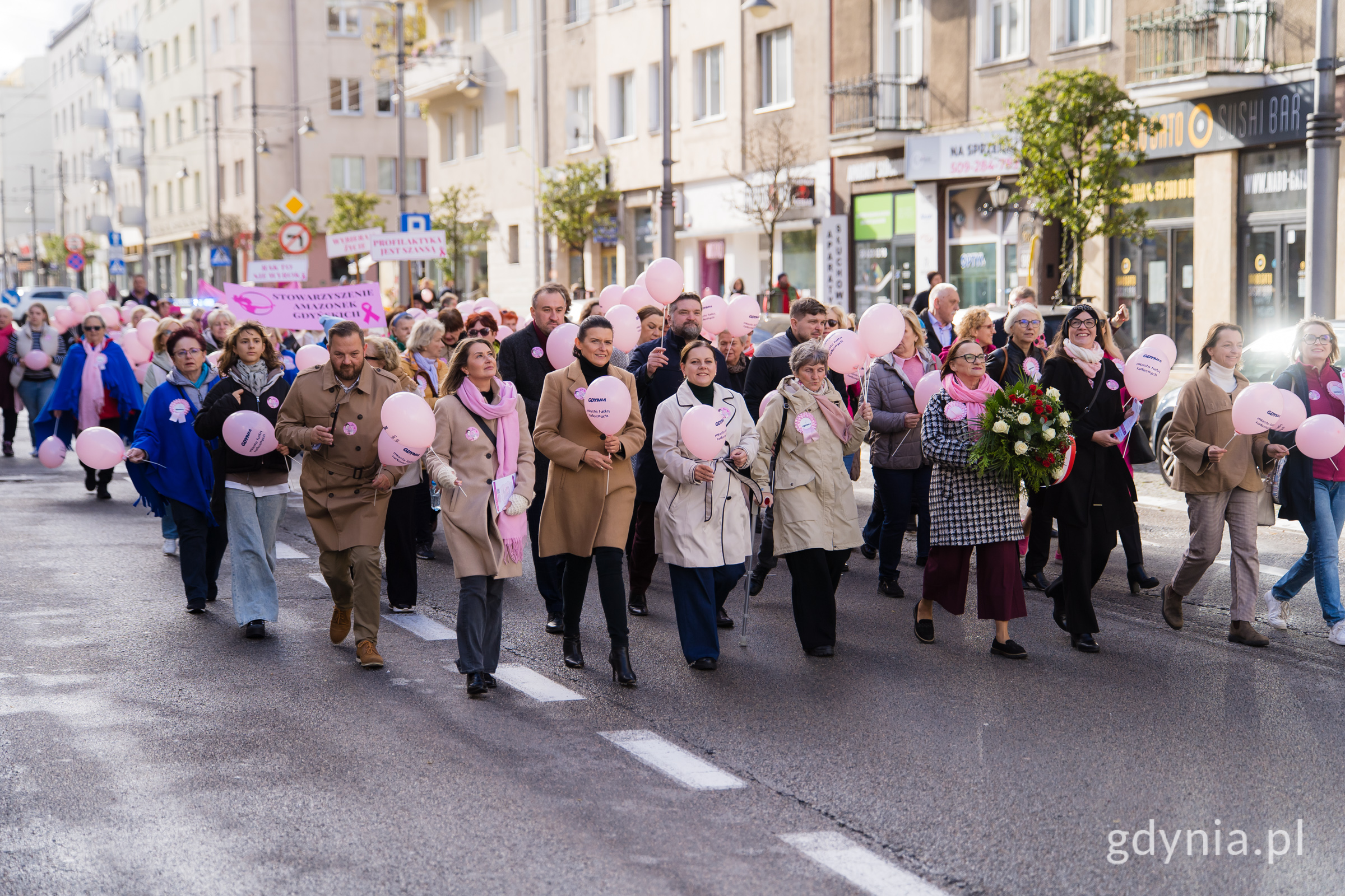 Uczestnicy Marszu Różowej Wstążki idący ul. Świętojańską. Wśród nich m.in. (od lewej): radni miasta Łukasz Strzałkowski i Monika Strzałkowska, wiceprezydent Gdyni Tomasz Augustyniak, Mariola Śrubarczyk-Cichowska - wiceprzewodnicząca Rady Miasta Gdyni, prezydent Gdyni Aleksandra Kosiorek, Tadeusz Szemiot - przewodniczący Rady Miasta Gdyni, Janina Łojba - prezes Stowarzyszenia Amazonek Gdyńskich, radni miasta Dominik Aziewicz, Beata Zastawna i Joanna Zielińska, posłanka Wioleta Tomczak, a także radne miasta Larysa Kramin oraz Agnieszka Tokarska 