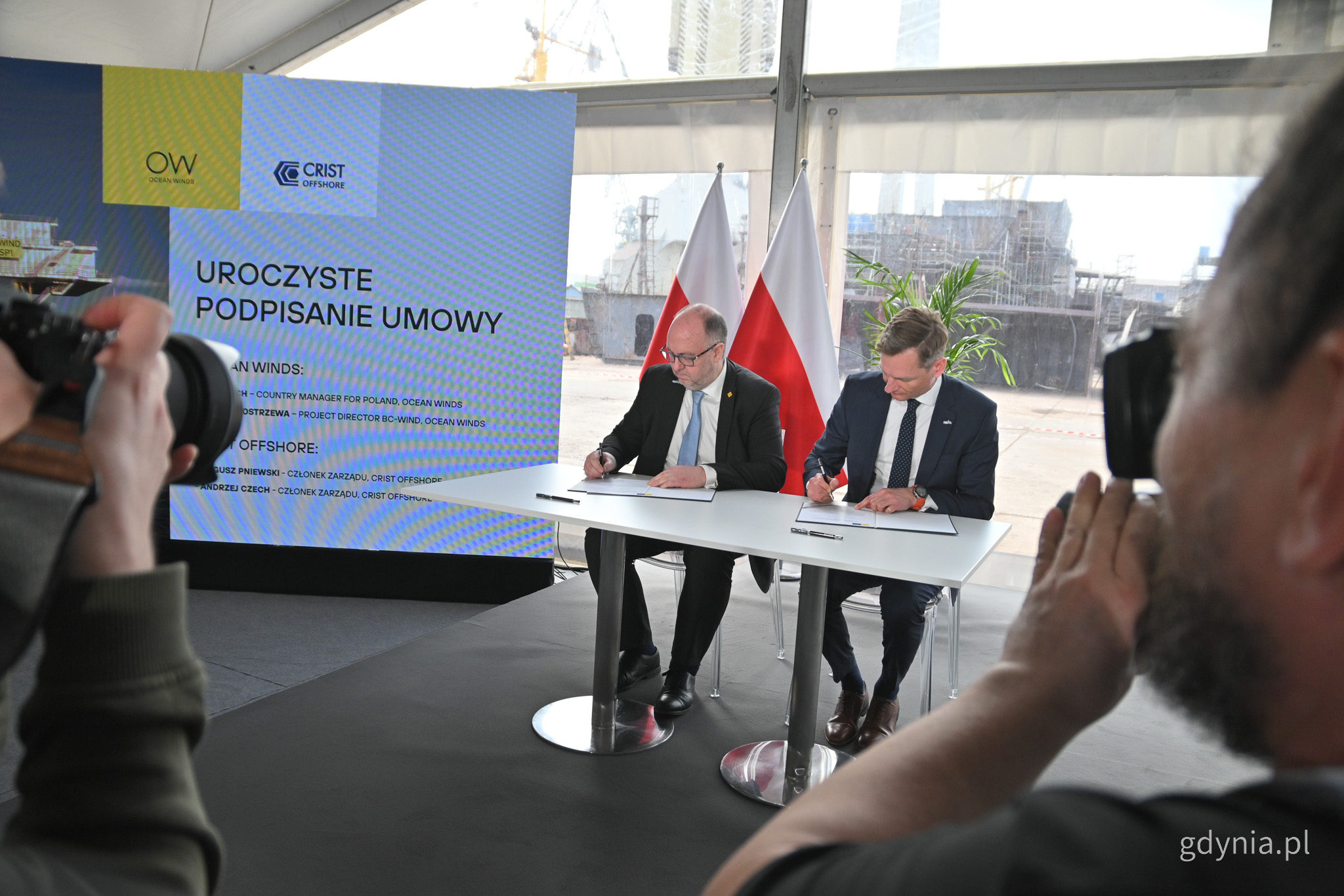 Namiot w stoczni Crist, dwóch mężczyzn w garniturach siedzi przy stole i podpisuje umowę, obok widoczni fotoreporterzy