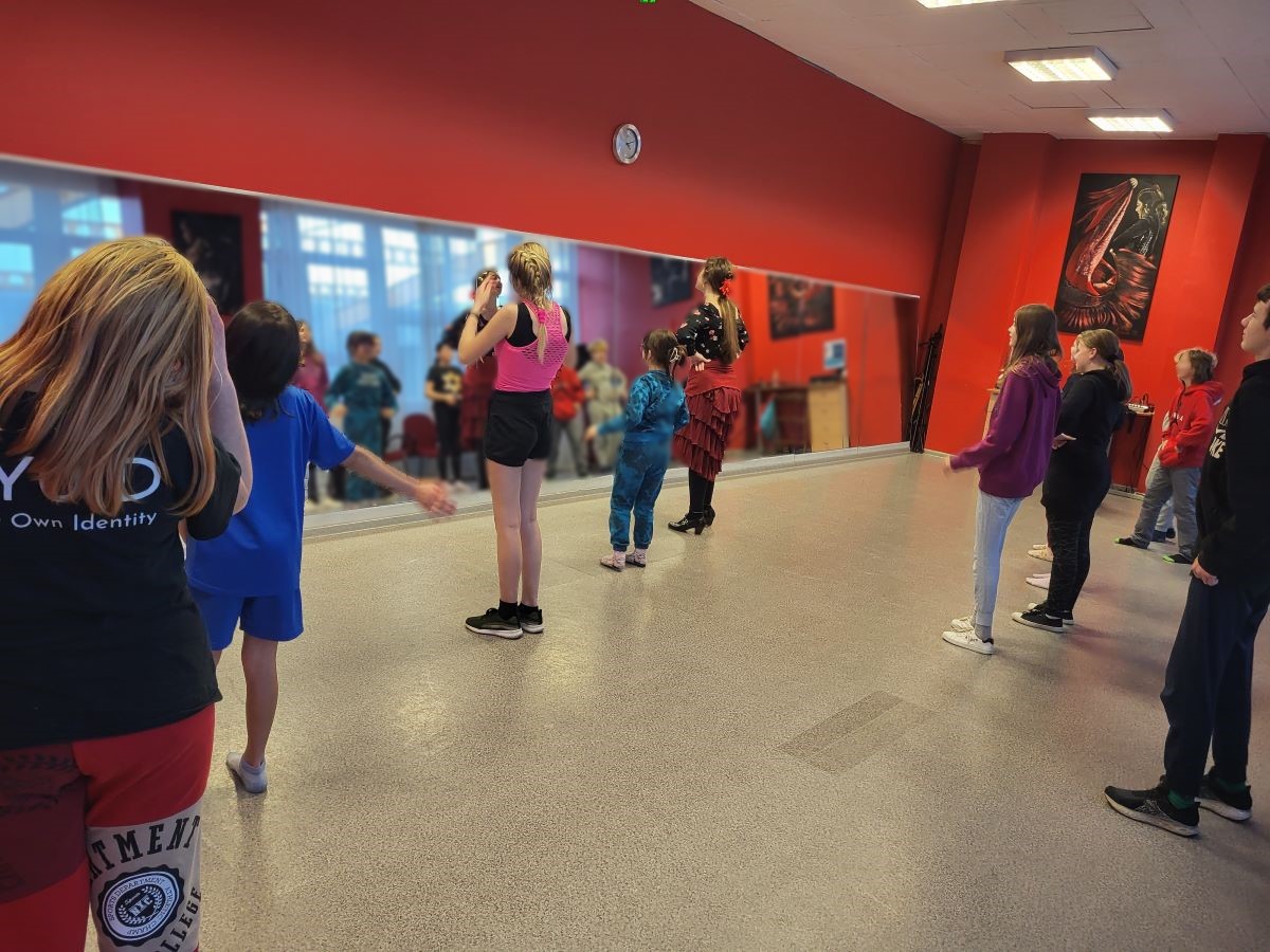 Zdjęcie przedstawia zajęcia taneczne będące elementem dnia hiszpańskiego w SPOT Światłowcy. Widzimy instruktorkę flamenco wraz z grupą dzieci. Grupa stoi przed lustrem ćwicząc ruchy taneczne.