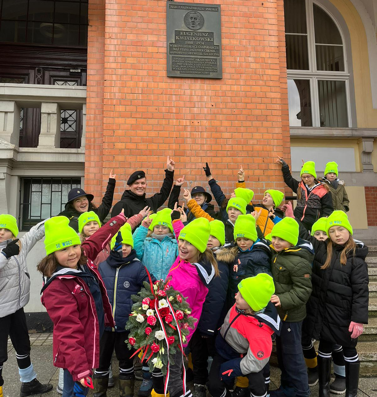 grupa harcerzy stoi pod tablicą pamiątkową i wskazuje na nią palcami.