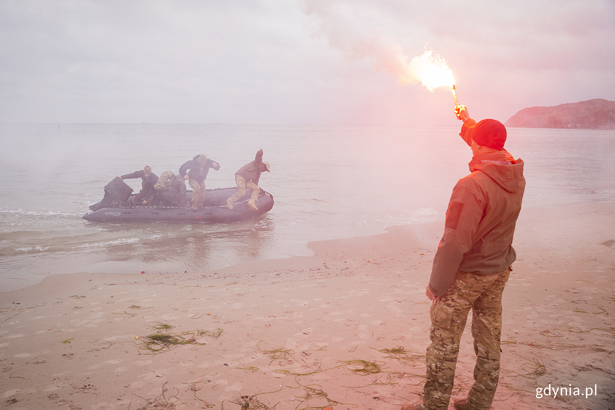 Na plaży stoi żołnierz z racą, z pontonu wyskakują komandosi.