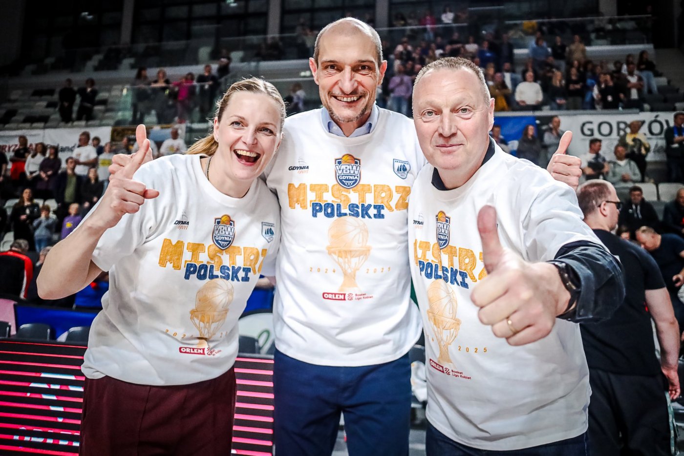 Hala sportowa, do zdjęć w białych koszulkach z napisami "Mistrz Polski" pozują trzy osoby: kobieta i dwóch mężczyzn, jeden z nich pokazuje uniesiony kciuk, w tle kibice na widowni