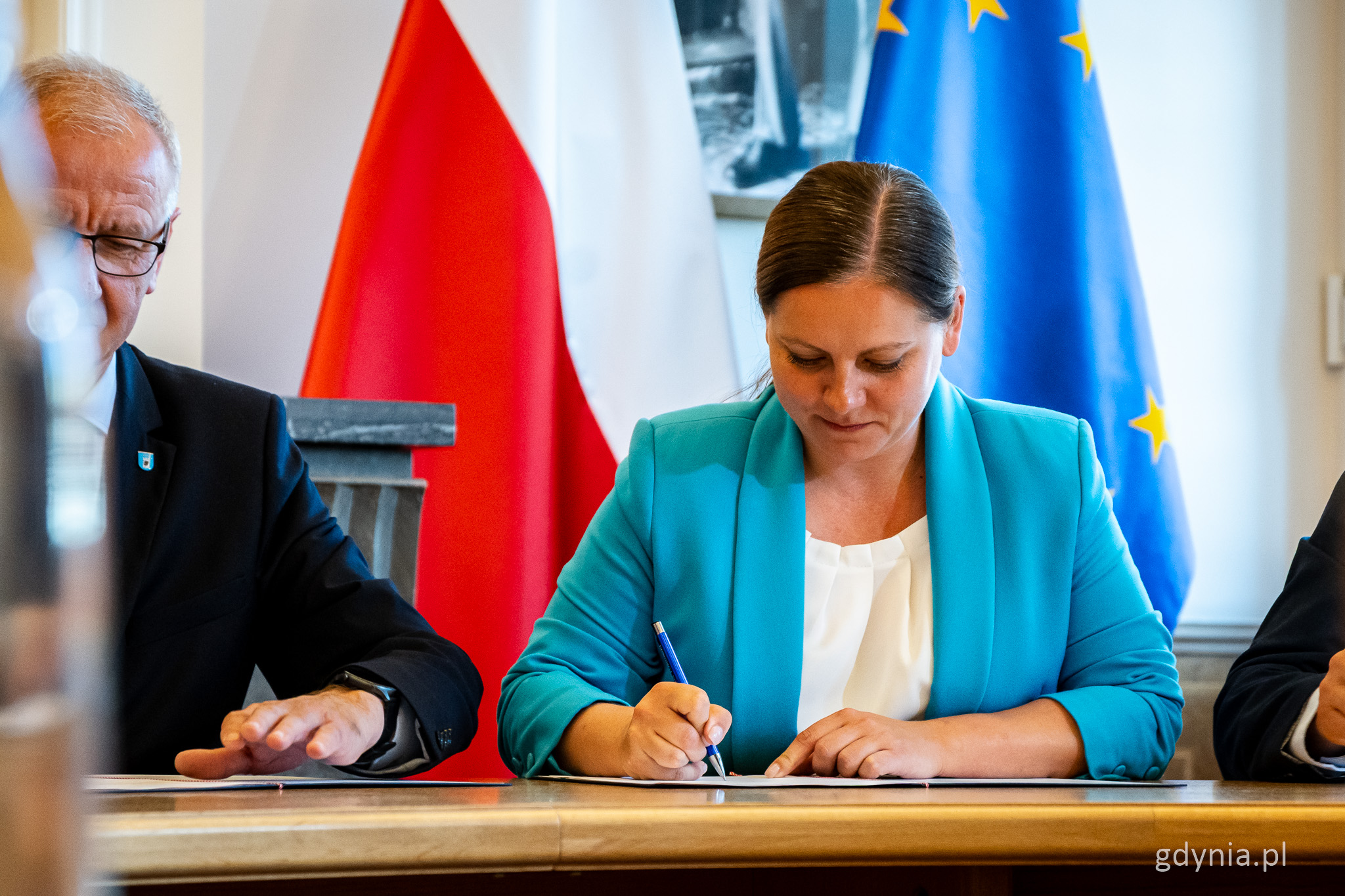 Sala w Pomorskim Urzędzie Wojewódzkim, przy stole dokumenty podpisuje Aleksandra Kosiorek, w tle flaga UE i Polski