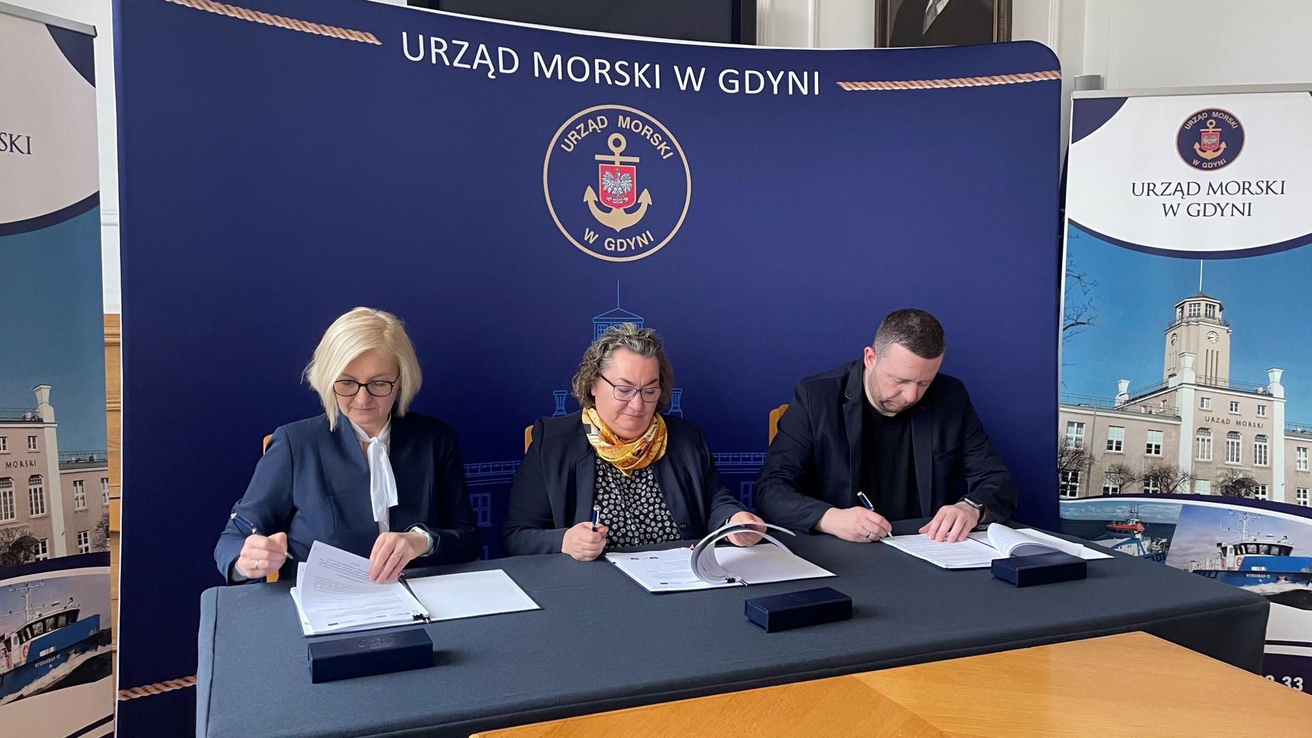 Pomieszczenie w Urzędzie Morskim, ścianka, stół, przy stole trzy osoby podpisują umowy. Siedzą Katarzyna Brzózka, Anna Stelmaszyk-Świerczyńska i Łukasz Porzuczek.