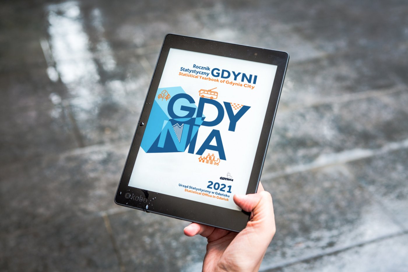 Ręka trzyma tablet na tle chodnika, na wyświetlaczu okładka Rocznika Statystycznego Gdyni z 2021 roku