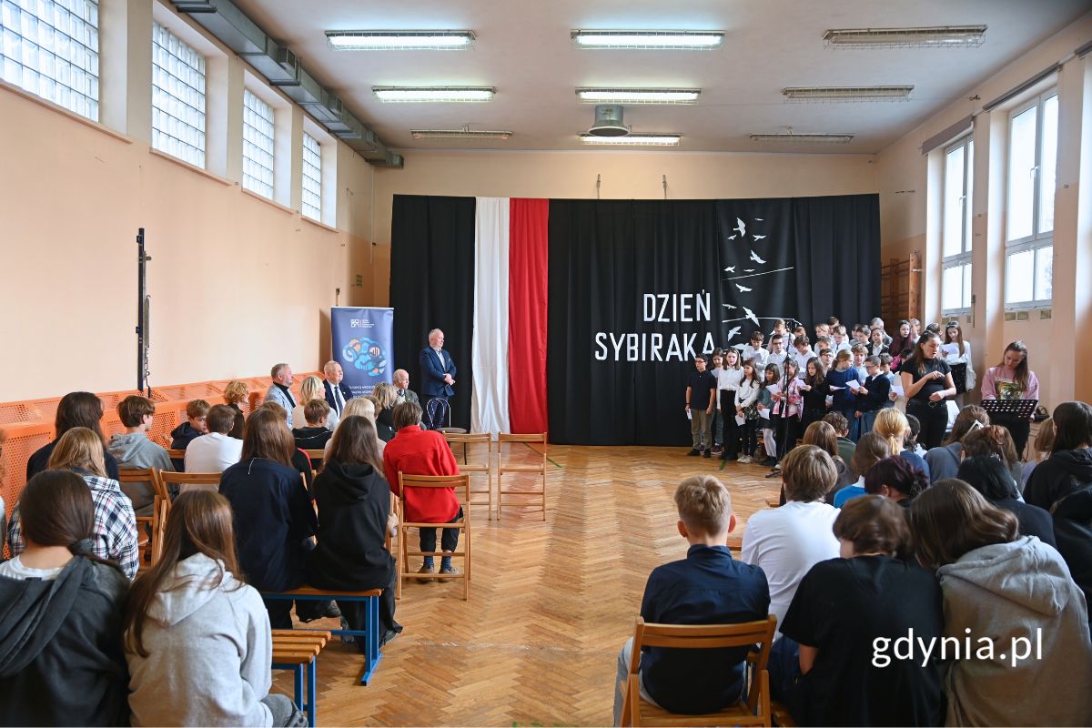 Uczniowie siedzący w ławkach na sali gimnastycznej tyłem do aparatu, w oddali widoczni seniorzy, chór dziecięcy oraz granatowa dekoracja z motywem polskiej flagi