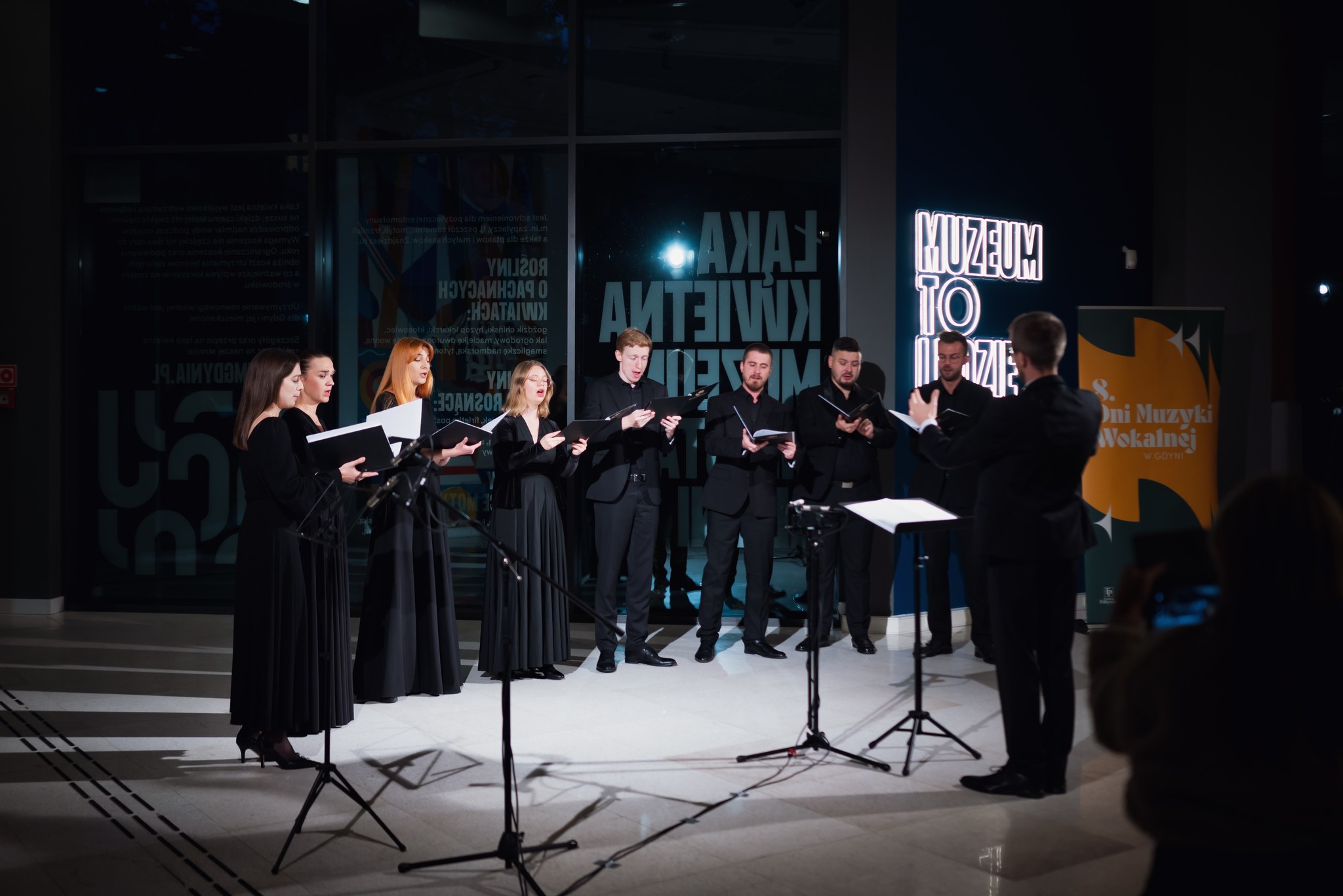 Hol w Muzeum Miasta Gdyni, chór śpiewa, obok dyrygent, ciemne pomieszczenie, podświetlony napis "Muzeum to ludzie"