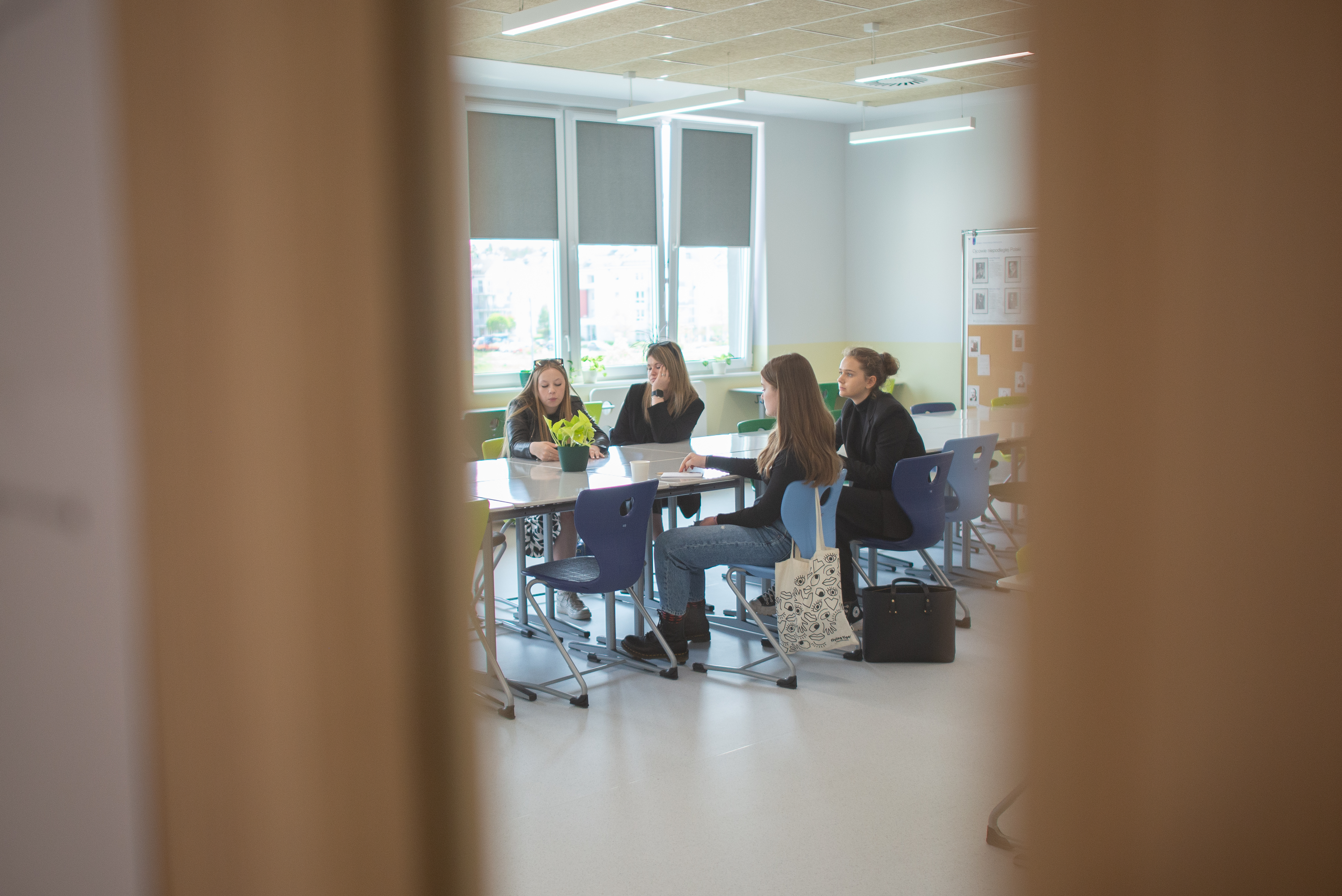 Przez wizjer w drzwiach widać grupę dziewcząt siedzącą w klasie przy stole