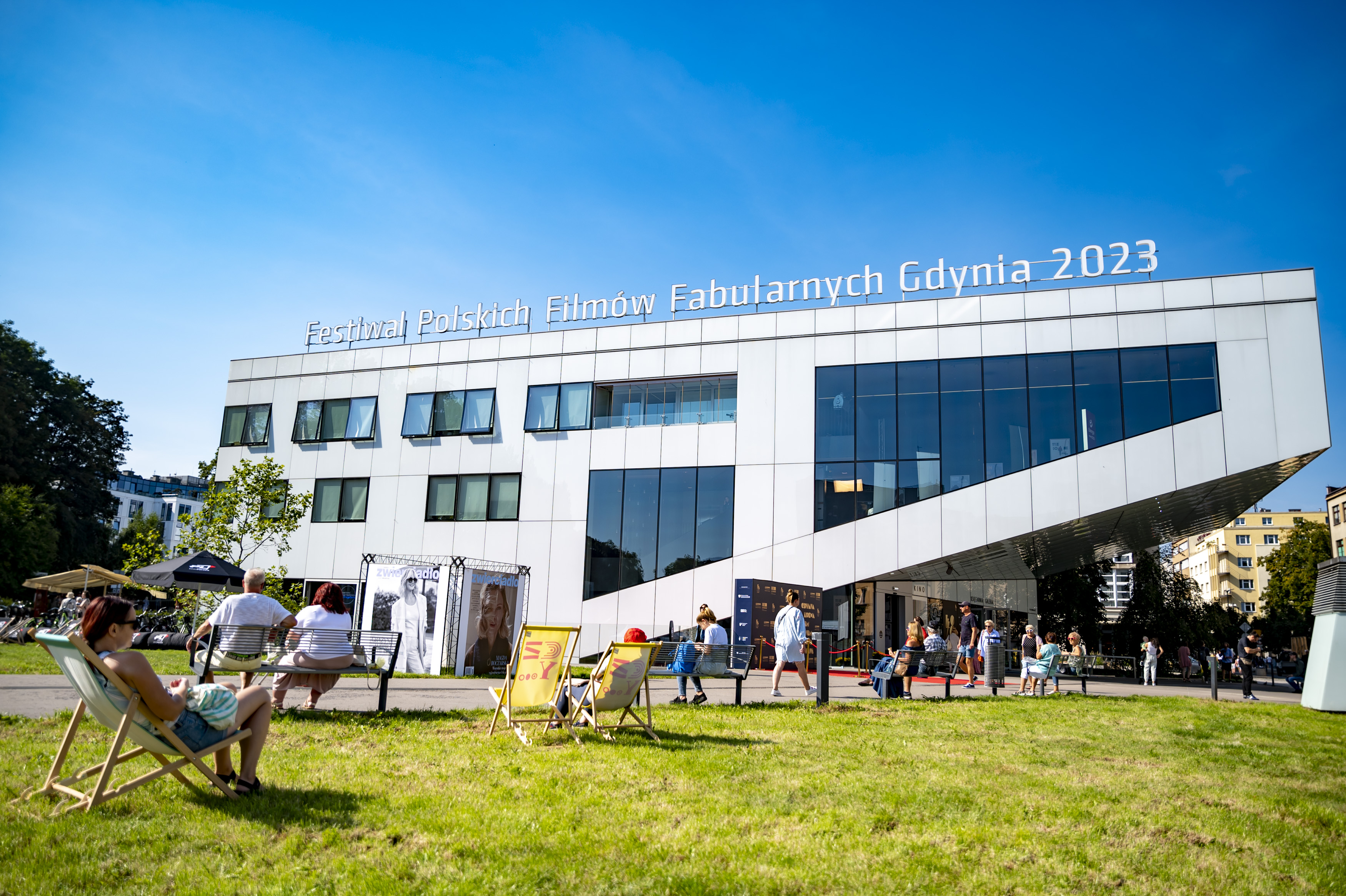 Gdyńskie Centrum Filmowe - budynek, plac Grunwaldzki, trawa, spacerujący ludzie, w tle budynki, słoneczny dzień