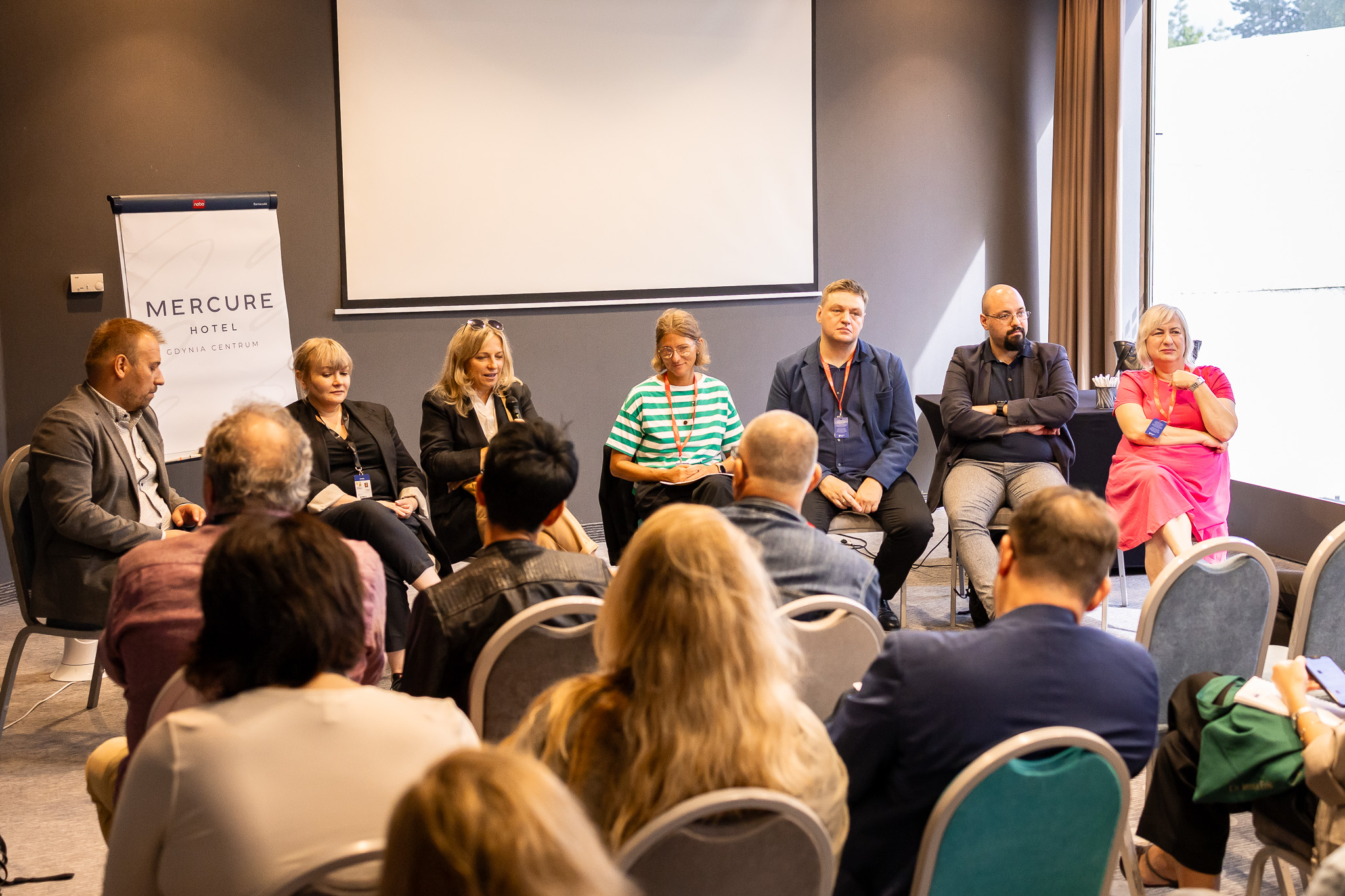 Sala konferencyjna, widownia pełna ludzi, na środku kilku rozmówców, ścianki z logotypami