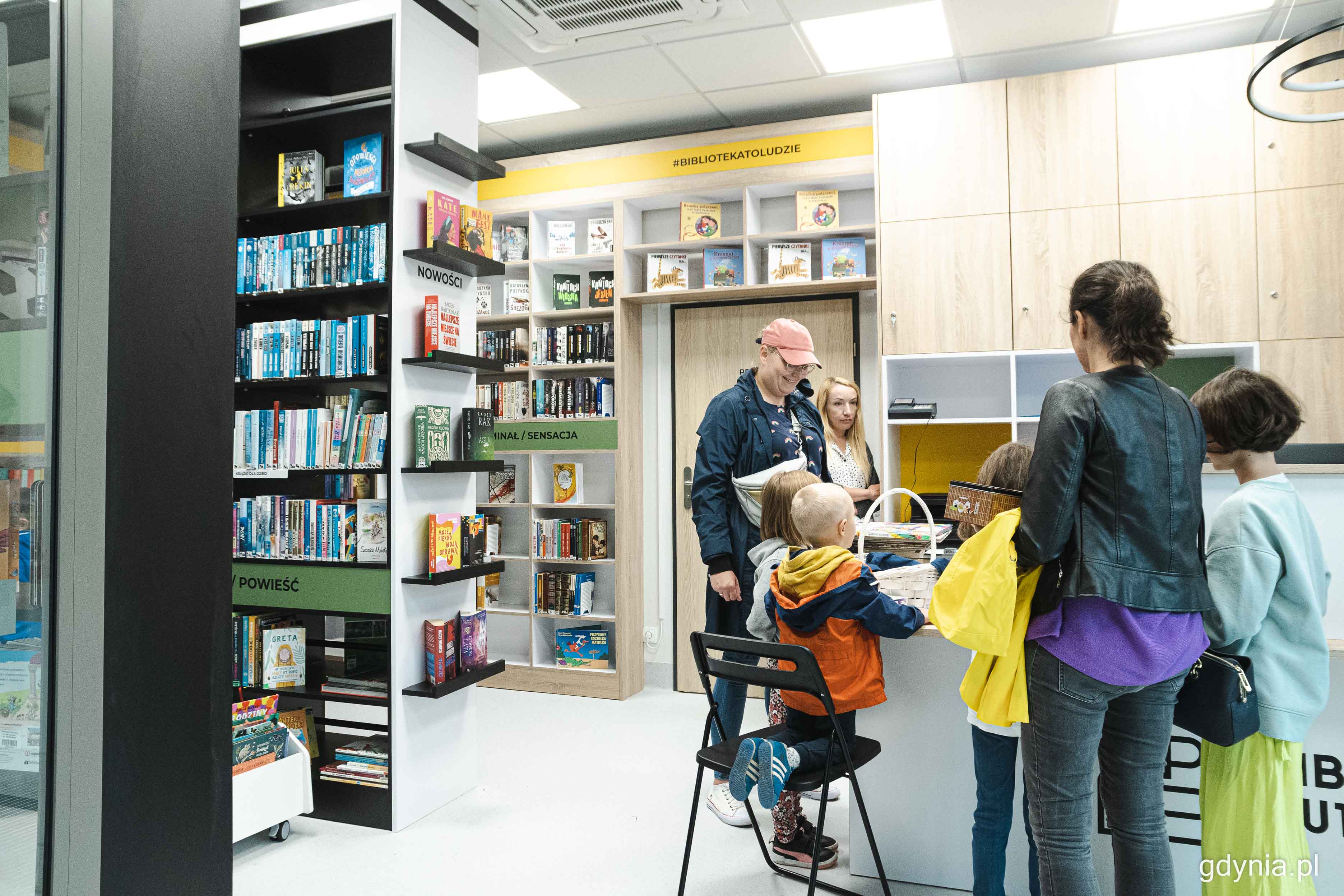 Gurpa osób stoi przy ladzie, Biblioteka Butikowa, książki na półkach i regałach