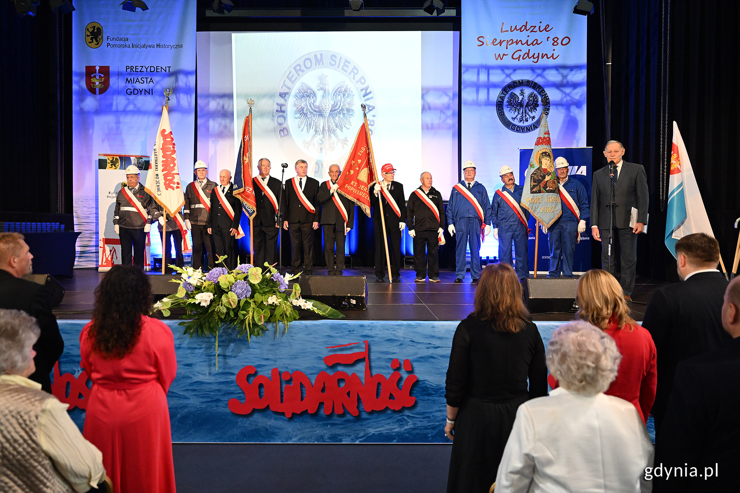 Scena w sali koncertowej Portu Gdynia, osoby na widowni, flagi Solidarności, na scenie grupa osób z pocztów sztandarowych