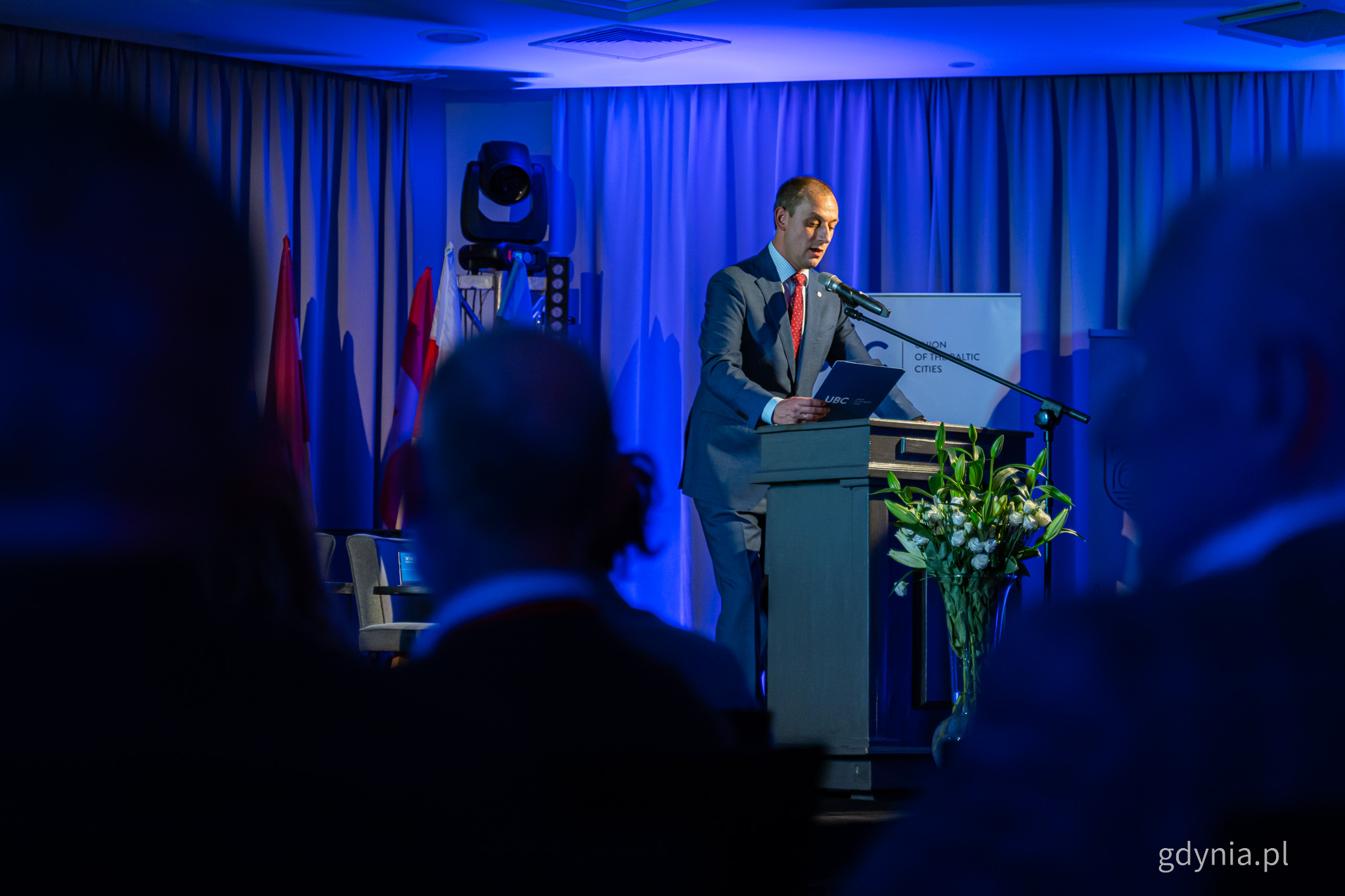 Sala konferencyjna, scena, niebieskie oświetlenie, przemawia przy mównicy Mantas Jurgutis, prezydent Związku Miast Bałtyckich