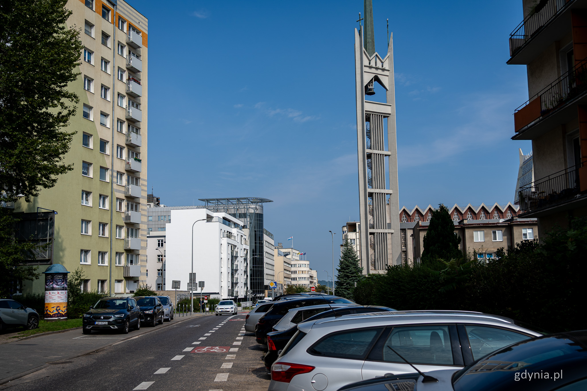 Ulica 3 Maja w Gdyni, samochody, jezdnia, bloki, kościół i biały budynek na drugim planie, widoczna biała ściana szczytowa