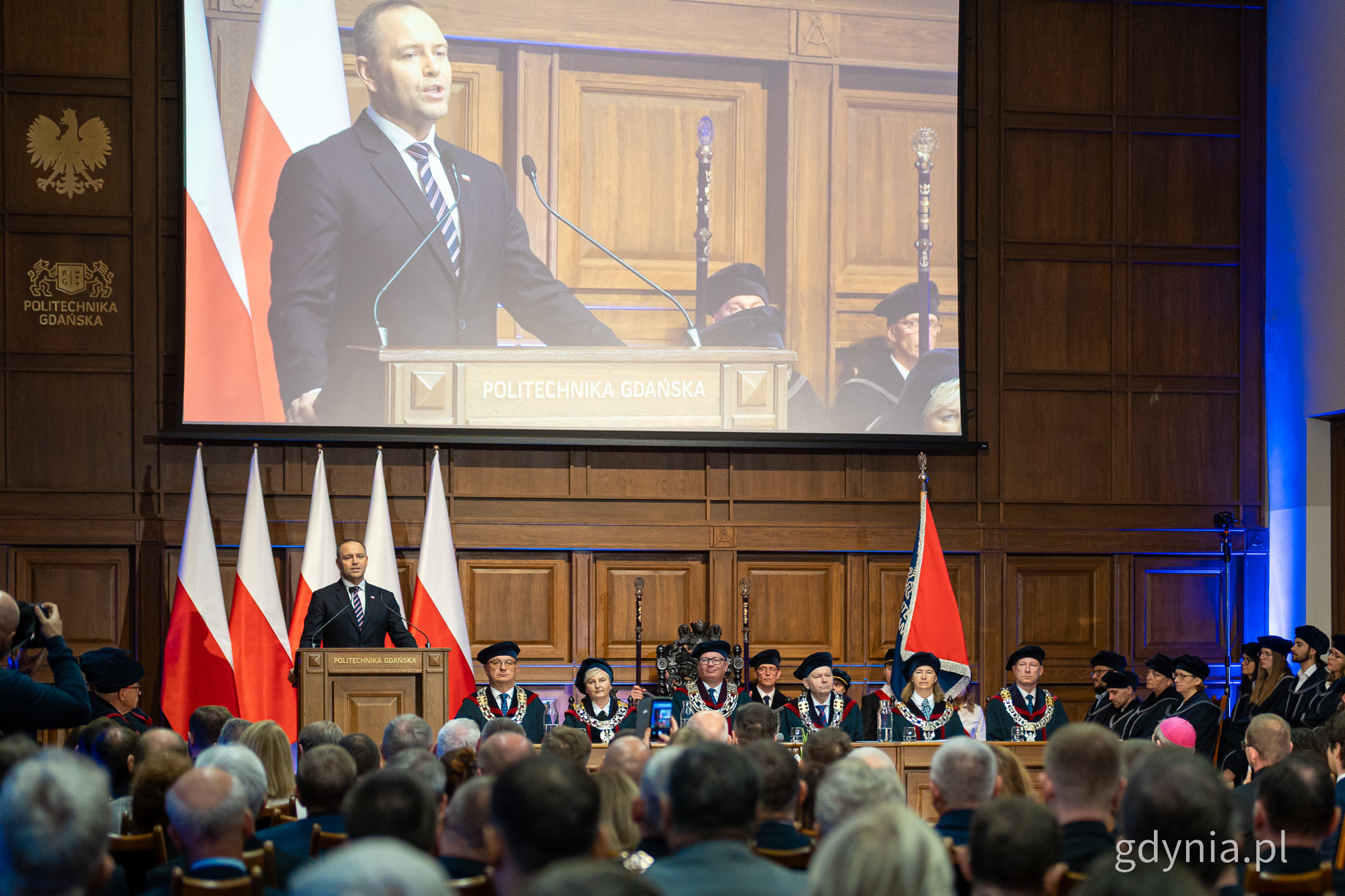 Aula PG, tłum osób, na mównicy Karol Nawrocki, nad nim ekran z transmisją przemówienia