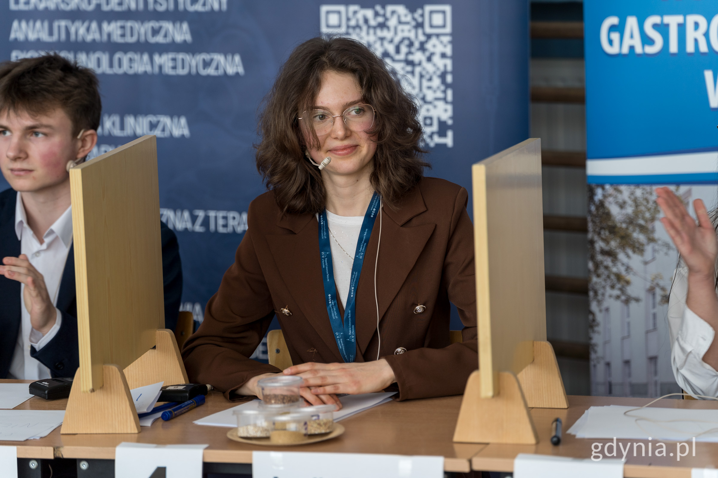 dziewczyna siedząca przy biurku, ma krecone brązowe włosy do ramion, okulary oraz mikrofon , jest  ubrana w brązową marynarkę, na stole przed nią stoją pojemniki z różnymi rodzajami kaszy, obok dziewczyny na sąsiednim stanowisku, siedzi chłopak 