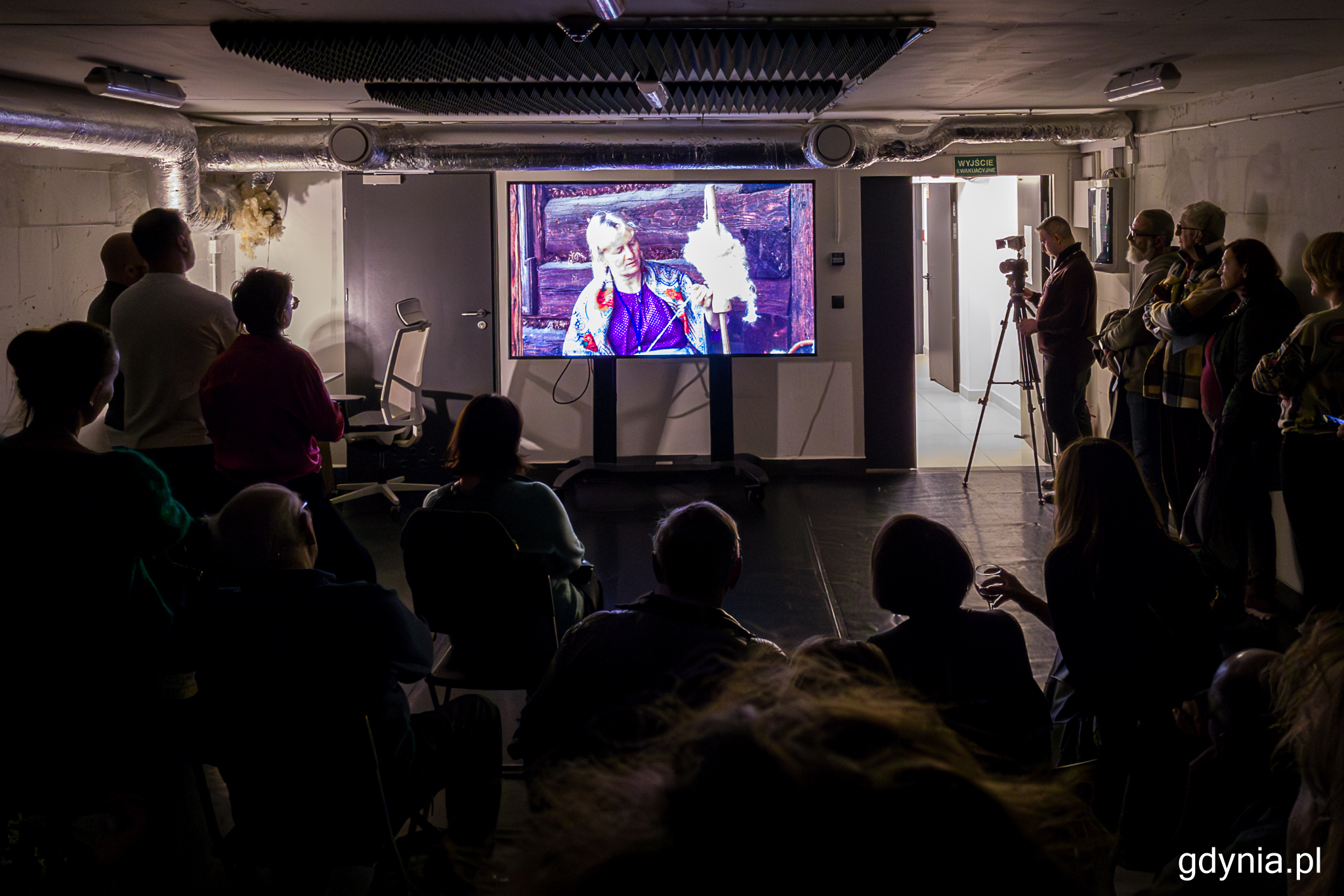 Sala, ekran z wyświetlanym filmem, półmrok, tłum ludzi dookoła ekranu, element wystawy "Część wspólna"