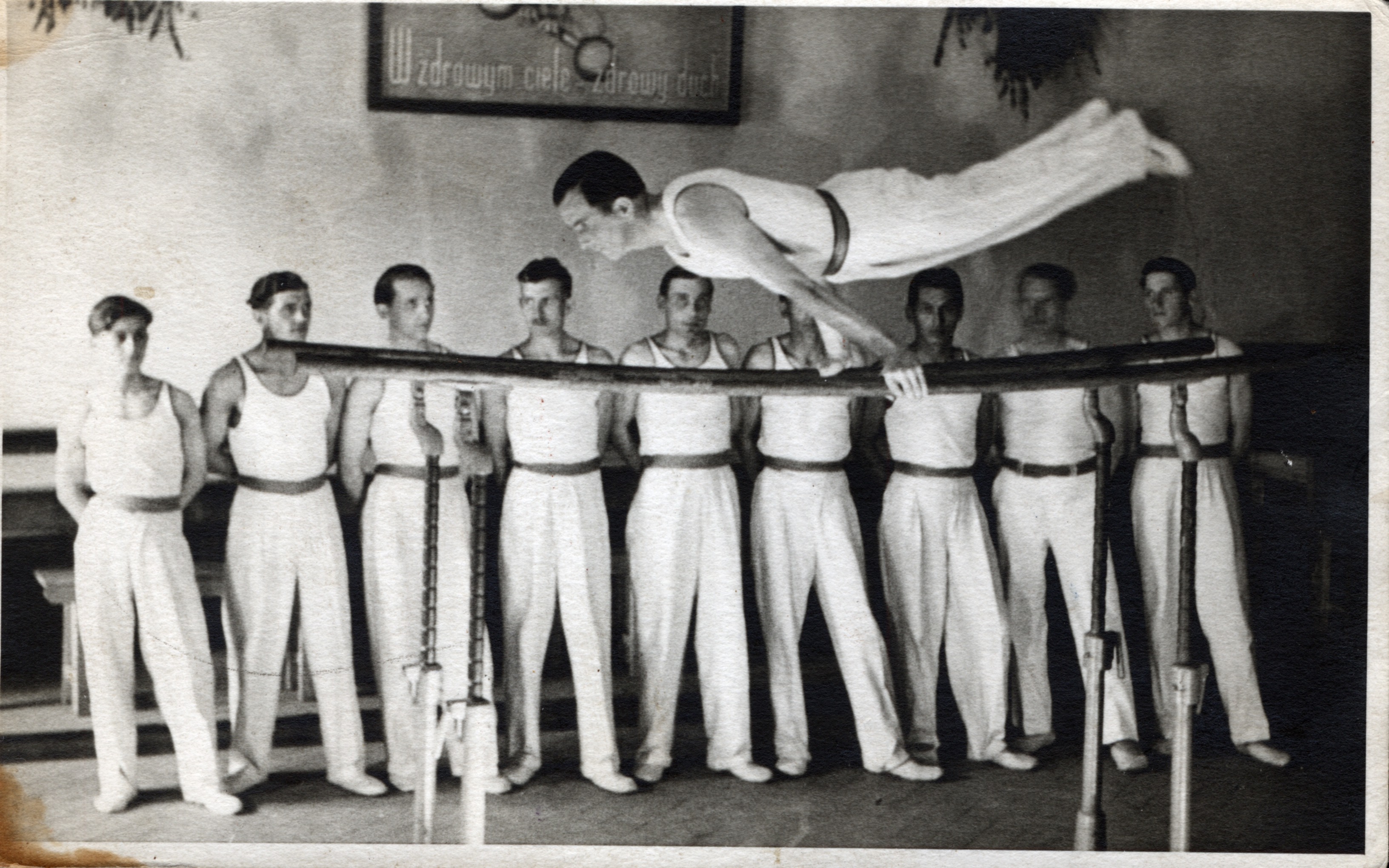 Czarno-białe zdjęcie z archiwum Muzeum Miasta Gdyni. Na zdjęciu widoczni są mężczyźni w białych strojach, którzy obserwują innego mężczyznę wykonującego akrobacje gimnastyczne na belkach.