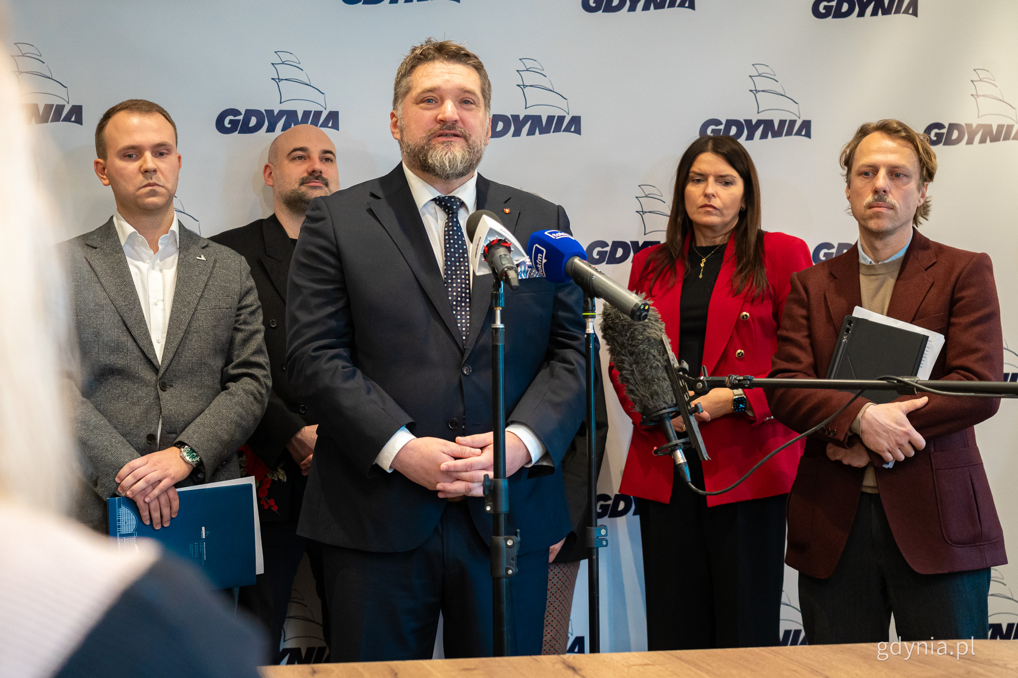 Sala w Urzędzie Miasta Gdyni, grupa radnych podczas konferencji prasowej, na środku przemawia do mikrofonów Tadeusz Szemiot, w tle logotypy Gdyni i białe tło