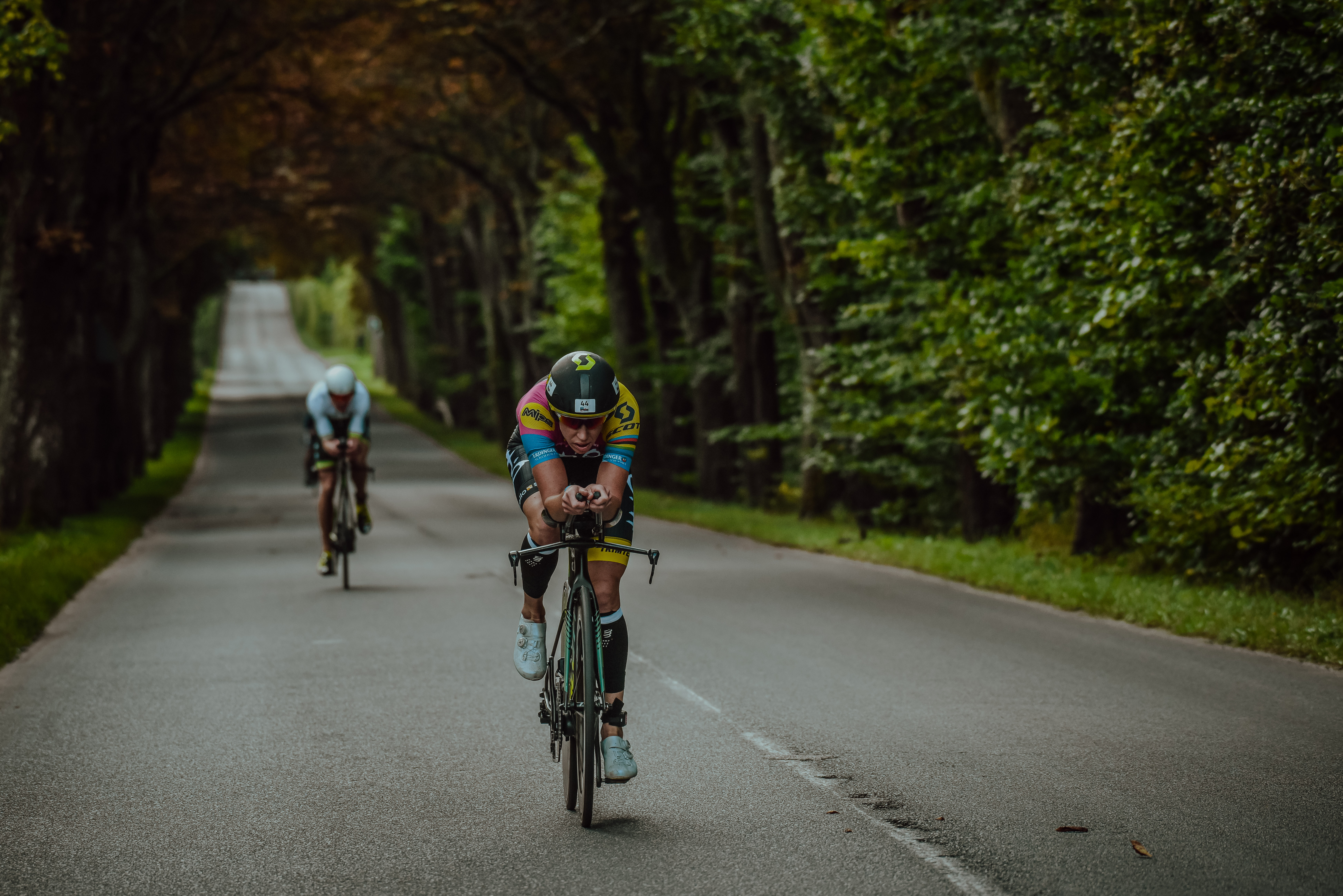 Uczestnicy zawodów triathlonowych jadą na rowerach po asfaltowej drodze otoczonej drzewami. Ręce opierają na kierownicy. Mają na sobie kolorowe stroje sportowe, okulary i kaski.