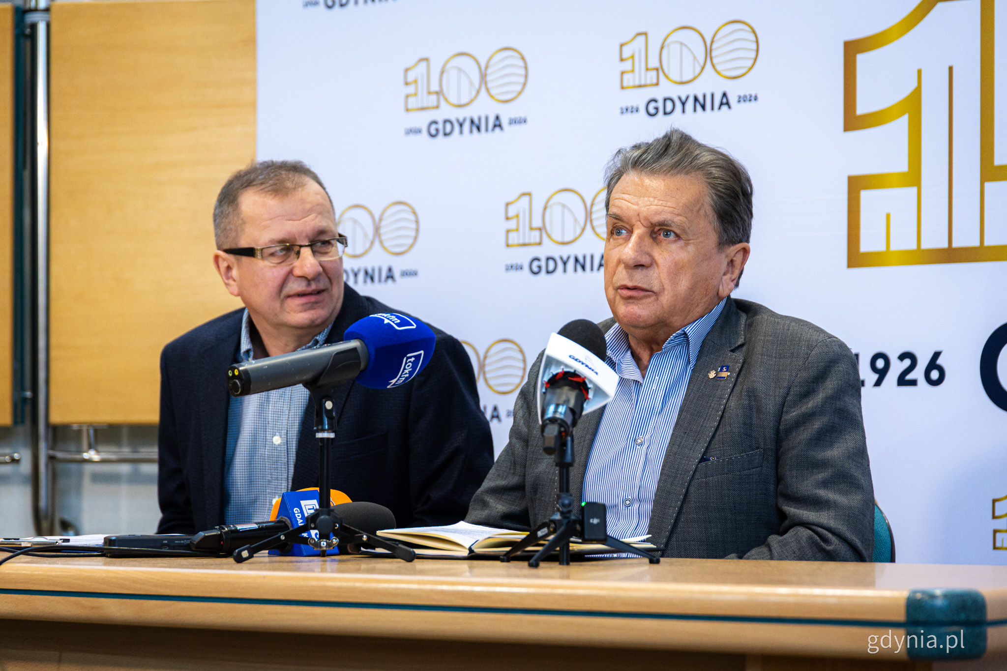 Dwaj mężczyźni siedzący przy stole i mający za plecami białą ściankę ze złotymi logotypami stulecia Gdyni