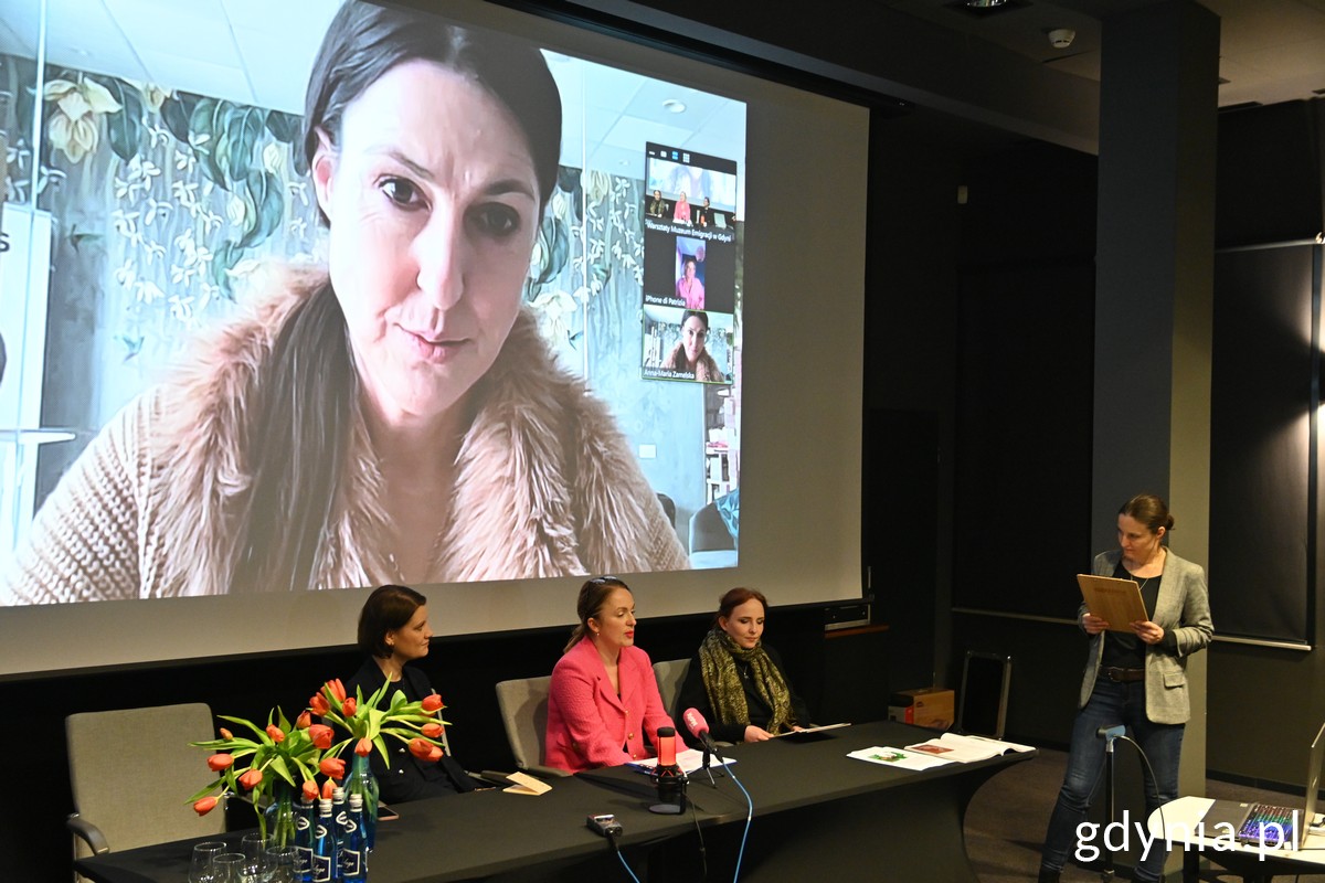 Trzy kobiety siedzą za stołem podczas konferencji, jedna kobieta stoi, na ekranie kolejna kobieta.