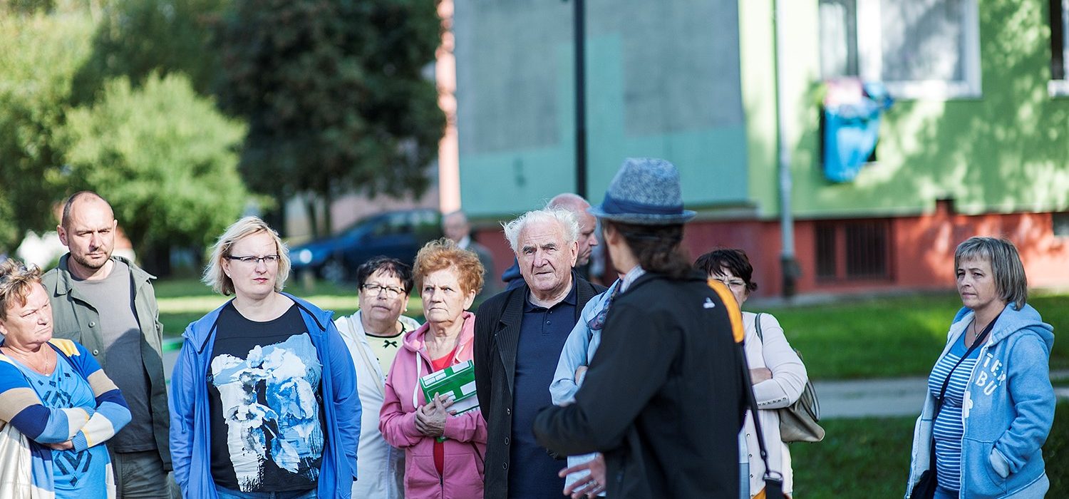 Grupa ludzi w różnym wieku (kobiety i mężczyźni). Znajdują się po środku osiedla. 