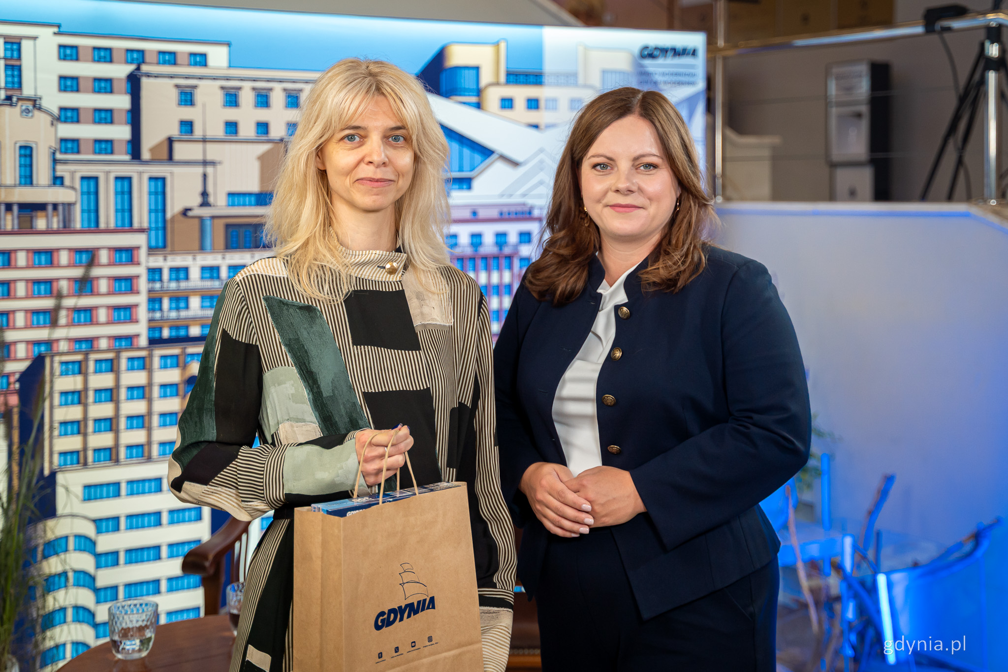 Joanna Łapińska i Aleksandra Kosiorek pozują do zdjęcia, za nimi meble, ścianka z ilustracjami modernistycznych budynk&oacute;w