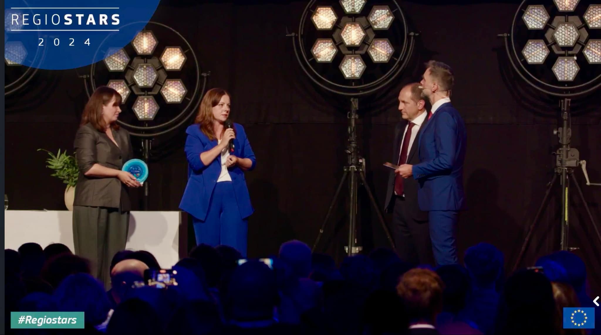 Grupa elegancko ubranych os&oacute;b na scenie, z kt&oacute;rych jednak przemawia do pozostałych