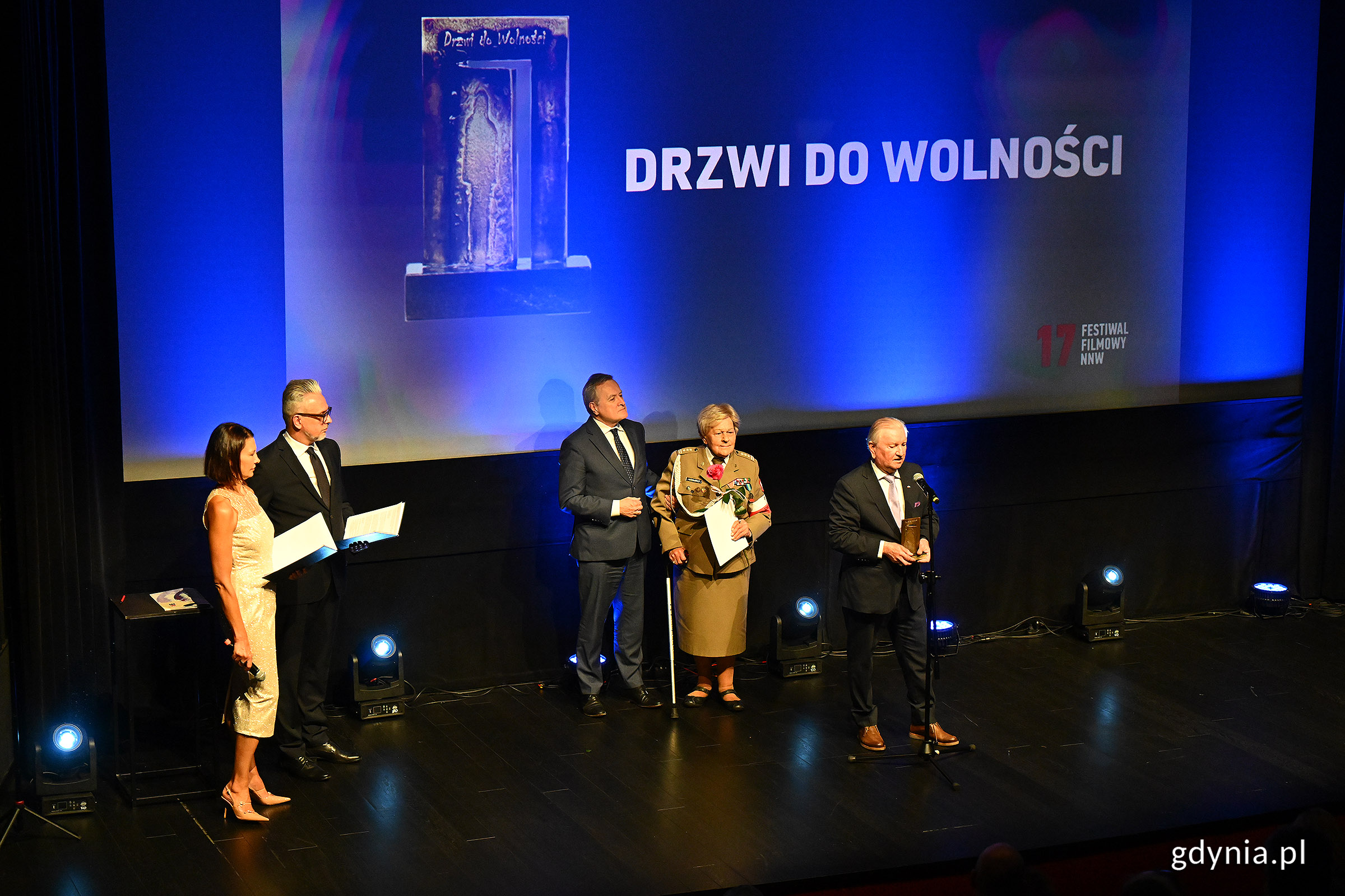Scena w sali Warszawa w GCF-ie, grupa os&oacute;b na scenie, napis na ekranie "Drzwi do wolności"
