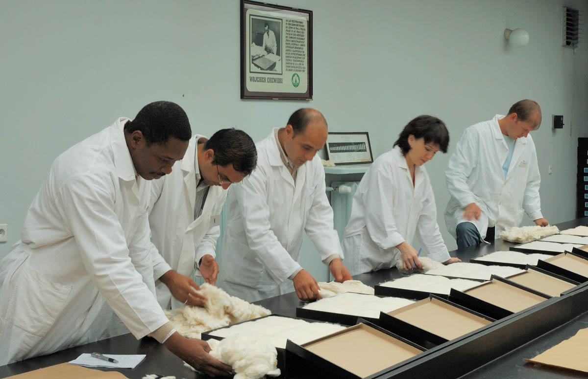 Sala, grupa os&oacute;b w białych fartuchach stoi przy stole, na stole zwoje bawełny