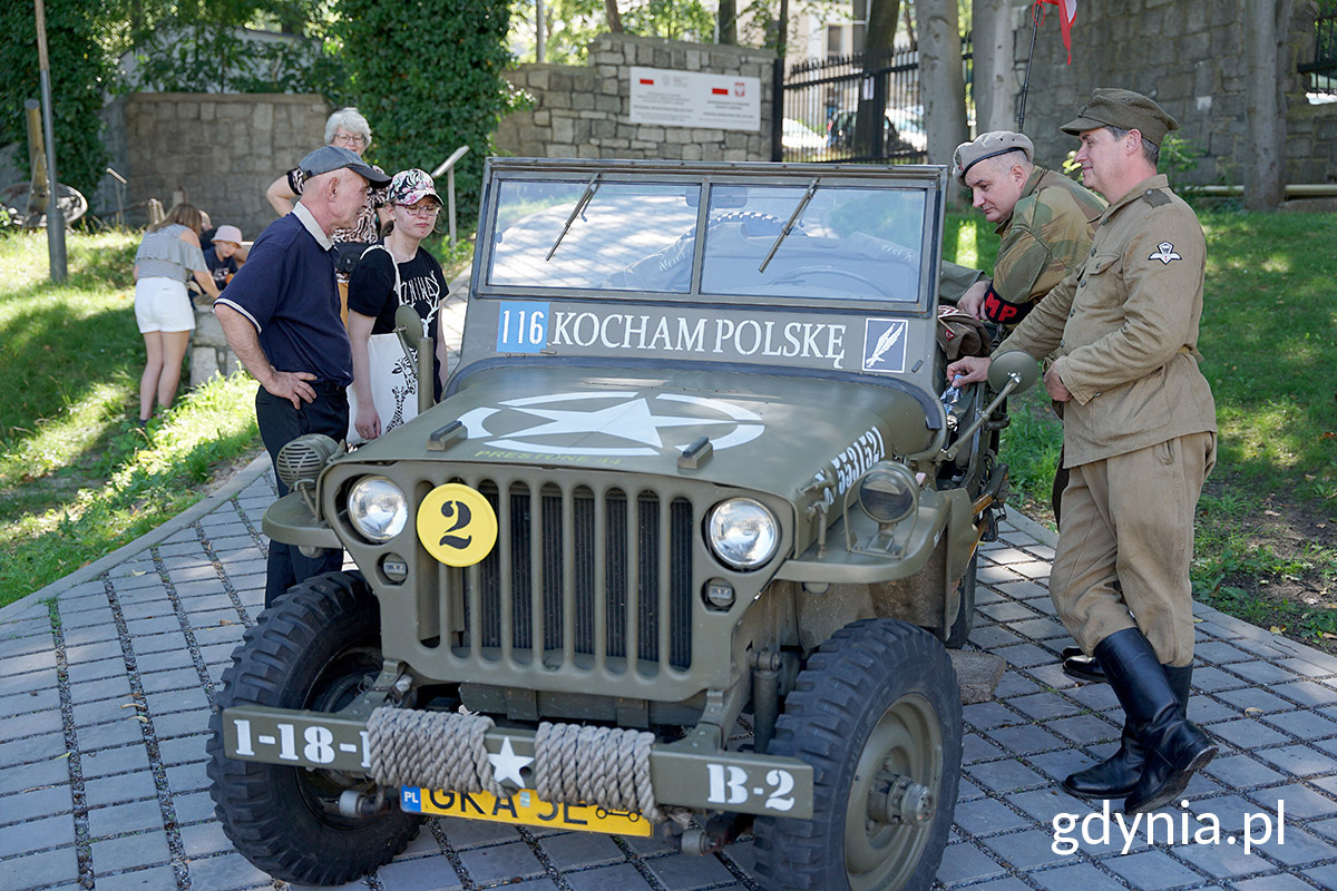 Wojskowy jeep z napisem 116, kocham Polskę. Obok (z lewej) stoi rodzina zwiedzająca Muzeum Marynarki Wojennej, z drugiej strony dw&oacute;ch mężczyzn w mundurach 