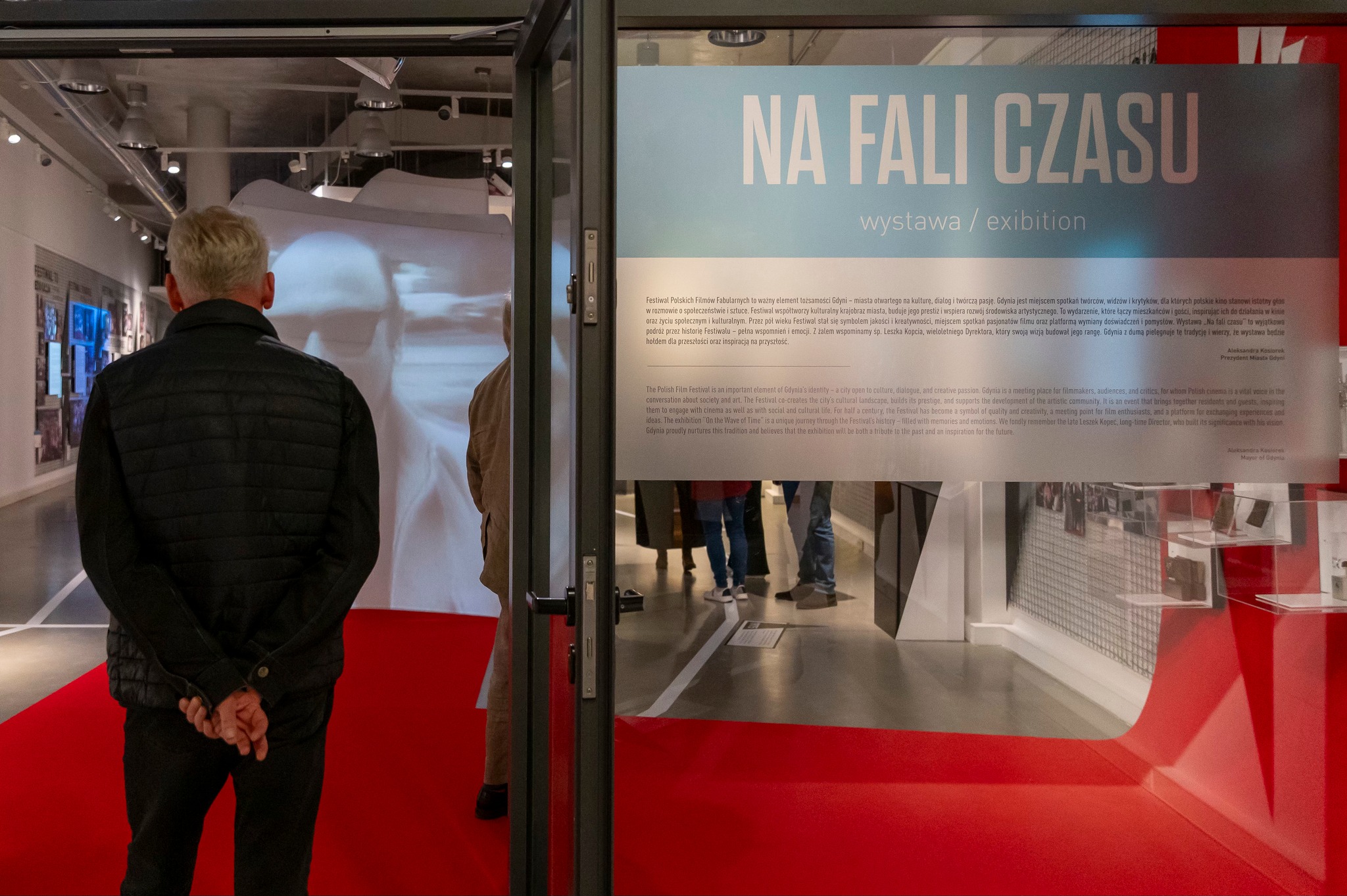 Wejście na wystawę "Na fali czasu" po lewej stronie jest mężczyzna zwiedzający wystawę, po prawej stronie plansza z nazwą wystawy. W tle pozostałe elementy wystawy